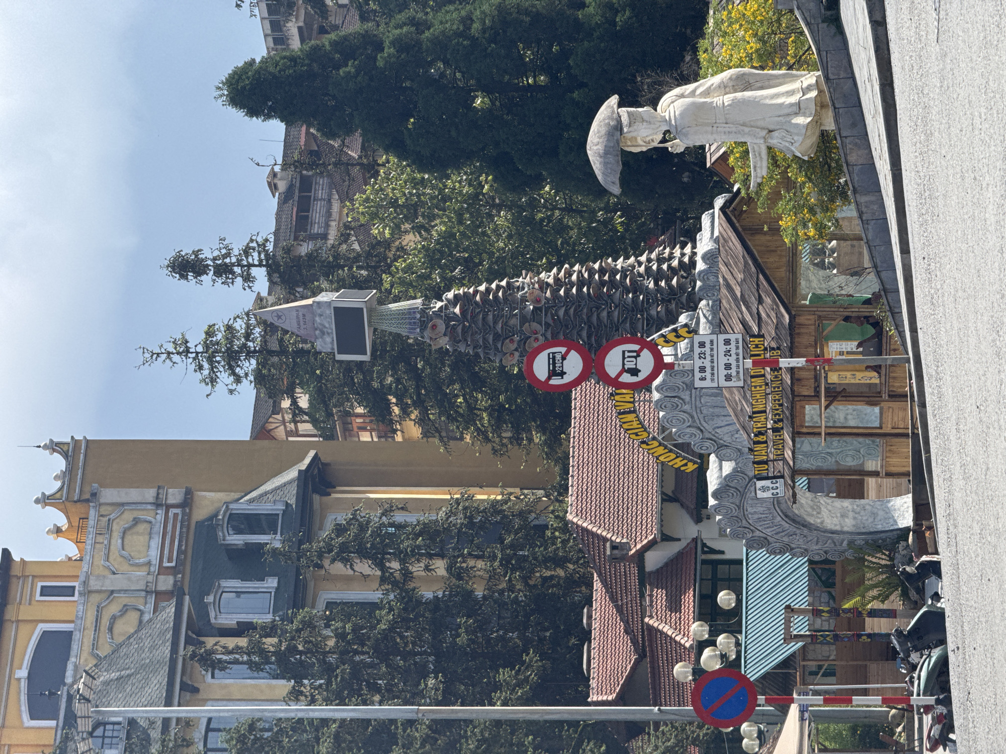This is a street scene in Sa Pa Ward, Vietnam, featuring a mix of architectural styles and decorative elements. In the foreground, there are several traffic signs including "No Entry" and "Weight Limit 10T", as well as a "No Parking" sign. A tall, conical structure rises, adorned with numerous small fans or reflectors, with a rectangular sign near the top. The buildings in the background exhibit a European style, with light yellow facades, gabled roofs, and dormer windows. Trees are interspersed among the buildings. A statue of a figure wearing a conical hat is visible in the upper right of the image. A decorated archway stands above the street, with the words "TƯ VĂN & TRẢI NGHIỆM DỊ LỊCH" written on it, alongside "CCC TRAVEL & EXPERIENCE." Motorbikes are parked on the street.