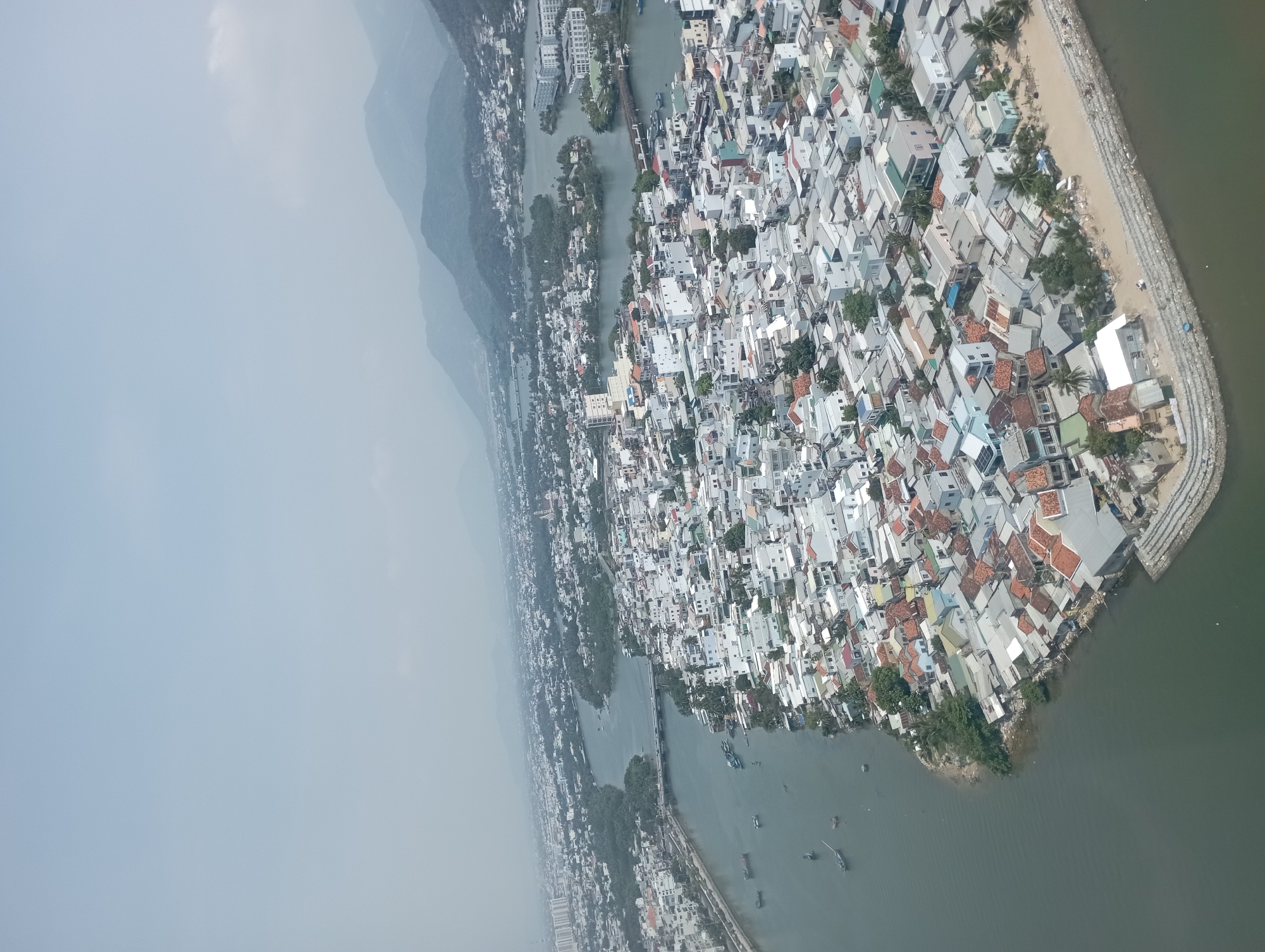 This is an aerial view of Nha Trang Ward, Vietnam. The scene depicts a densely populated coastal area. Numerous houses with predominantly white and grey roofs are interspersed with patches of greenery. A small river cuts through the city, with a mountain range visible in the background. The river meets the sea at the bottom right of the image. A barrier constructed of large stones lines the coast. Several boats are visible on the river. The image is somewhat hazy.
