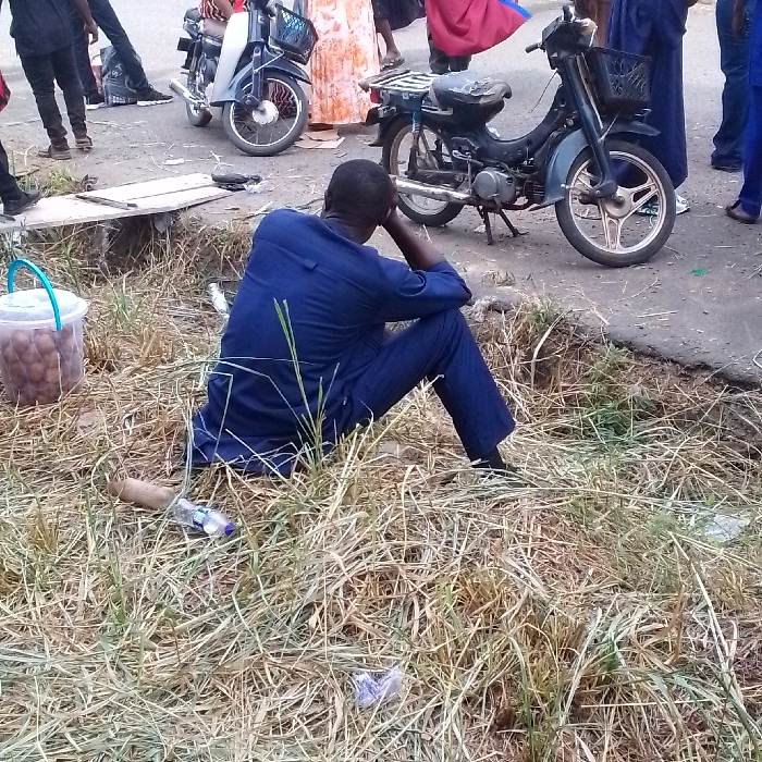The scene in Umueshi, Nigeria, depicts an outdoor area adjacent to a paved road. A dark-skinned male, wearing a dark blue long-sleeved shirt and matching trousers, sits on dry, yellowish-green grass, facing away from the viewer. His left elbow rests on his left knee, and his left hand supports his head. A clear plastic bucket with a light blue handle, filled with numerous brown spherical objects, is positioned to his left on the grass. A discarded plastic water bottle and a cylindrical object lie nearby. On the paved road, two motorcycles are present: a silver-grey model with a front basket on the left, and a dark blue-green model with a front basket and a rear rack on the right, resting on its kickstand. Multiple individuals, partially visible and dressed in various clothing including black, orange-patterned, red, and blue, are standing or moving in the background on the road. The immediate ground consists of sparse, dry grass with some litter.
