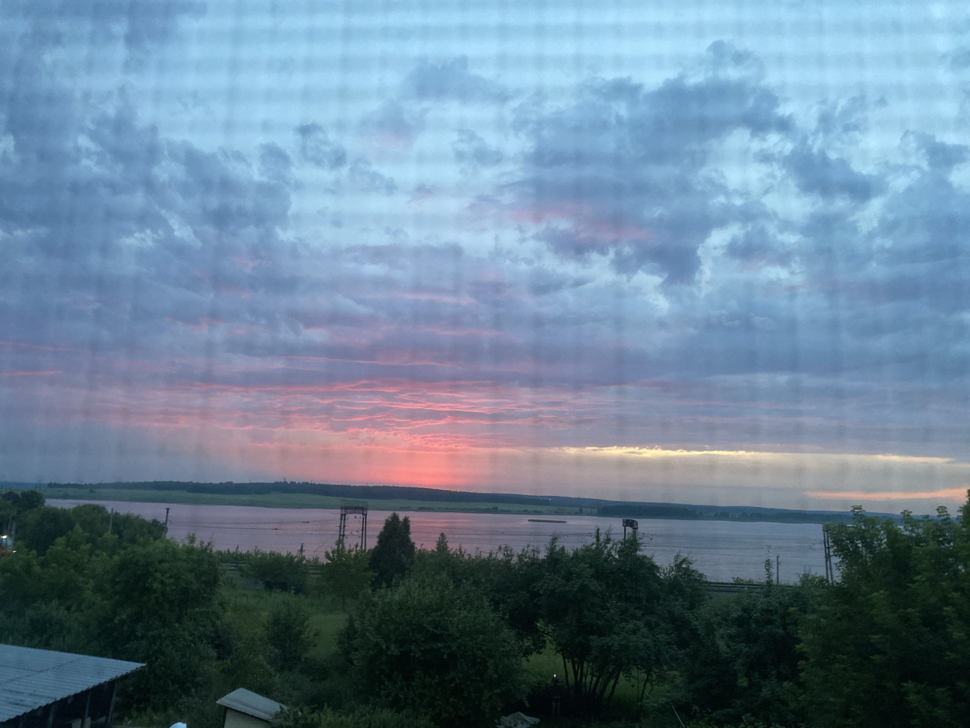 The image depicts a wide river at sunset, seen through a grid-like pattern, possibly a window screen or blinds. The sky is filled with clouds, with streaks of pink and orange near the horizon where the sun is setting. The river is calm and reflects the colors of the sky. On the far bank of the river, there is a flat landscape with some trees and what appear to be industrial structures. In the foreground, there is a dense line of trees and vegetation, with some man-made structures visible, including a roof with corrugated metal. The overall atmosphere is serene and tranquil, capturing the beauty of a natural landscape at dusk. The location is stated to be in the city of Smolino, Russia.