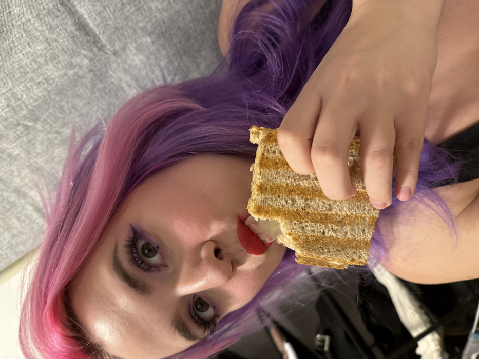 A person with pink and purple hair holds a sandwich to their lips. The sandwich is partially eaten, revealing the filling. The person has fair skin, dramatic purple eye makeup, and red lipstick. The background is a mix of neutral grey and glossy black, suggesting a casual or intimate setting. The image is rotated, giving a tilted perspective of the subject.