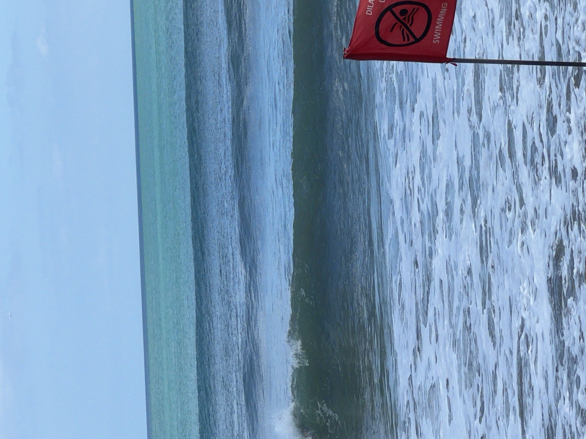 A red flag with a "no swimming" symbol and text in Indonesian is planted in the ocean near Kuta, Indonesia. The flag is attached to a grey pole and is partially submerged in the foamy water. The ocean is rough with large waves, and the sky is a pale blue with a few clouds. The scene depicts a warning against swimming due to hazardous conditions in the water.