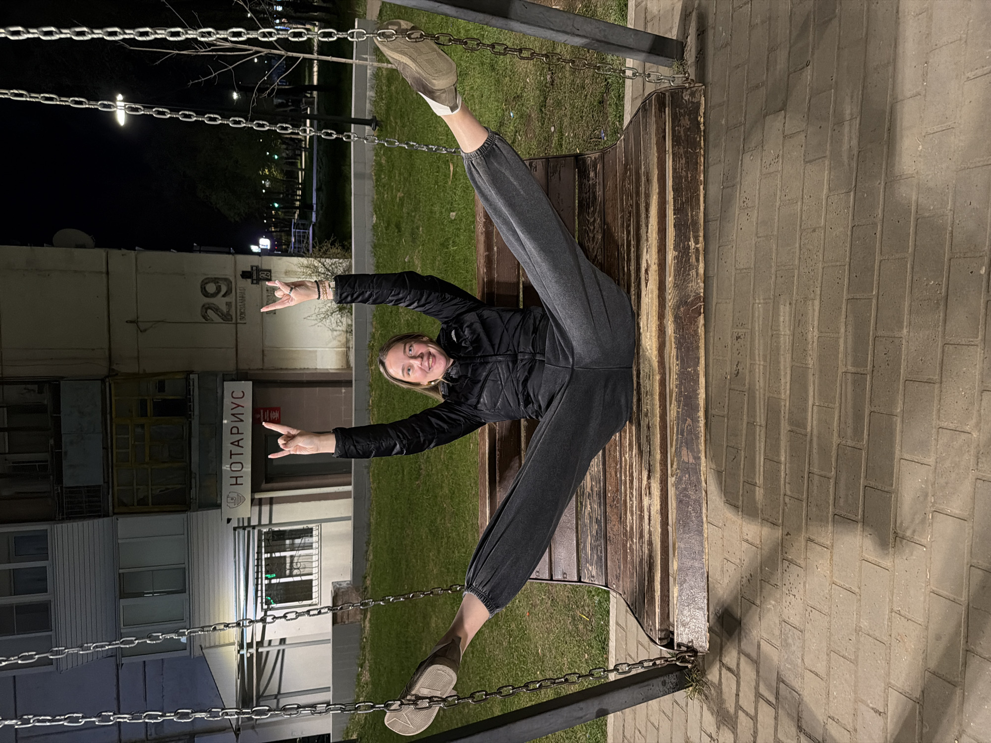 A woman is sitting on a swing in Chaikovsky, Russia, at night. She is wearing a black jacket and gray sweatpants, with khaki shoes. She is making a "rock on" hand gesture with both hands. The building behind her has a sign that reads "НОТАРИУС" (Notary) and the number 29. The area around the swing is paved with stone bricks, and there is a patch of grass in the background.