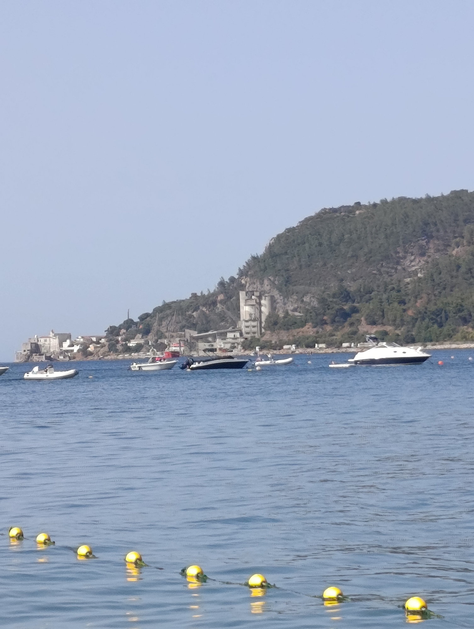 The image depicts a coastal scene, likely in Setúbal, Portugal, with a prominent hillside covered in dense green vegetation in the background. Several structures, including buildings and what appears to be a cement plant, are situated along the lower slopes of the hill. The foreground is dominated by a body of blue water, possibly a bay or harbor. A series of yellow buoys are floating in a line across the lower portion of the frame. Several boats are visible in the water, including a white speedboat, a smaller white boat, and a dark-colored vessel further out. The sky is clear and light blue. The overall impression is one of a sunny day by the sea.