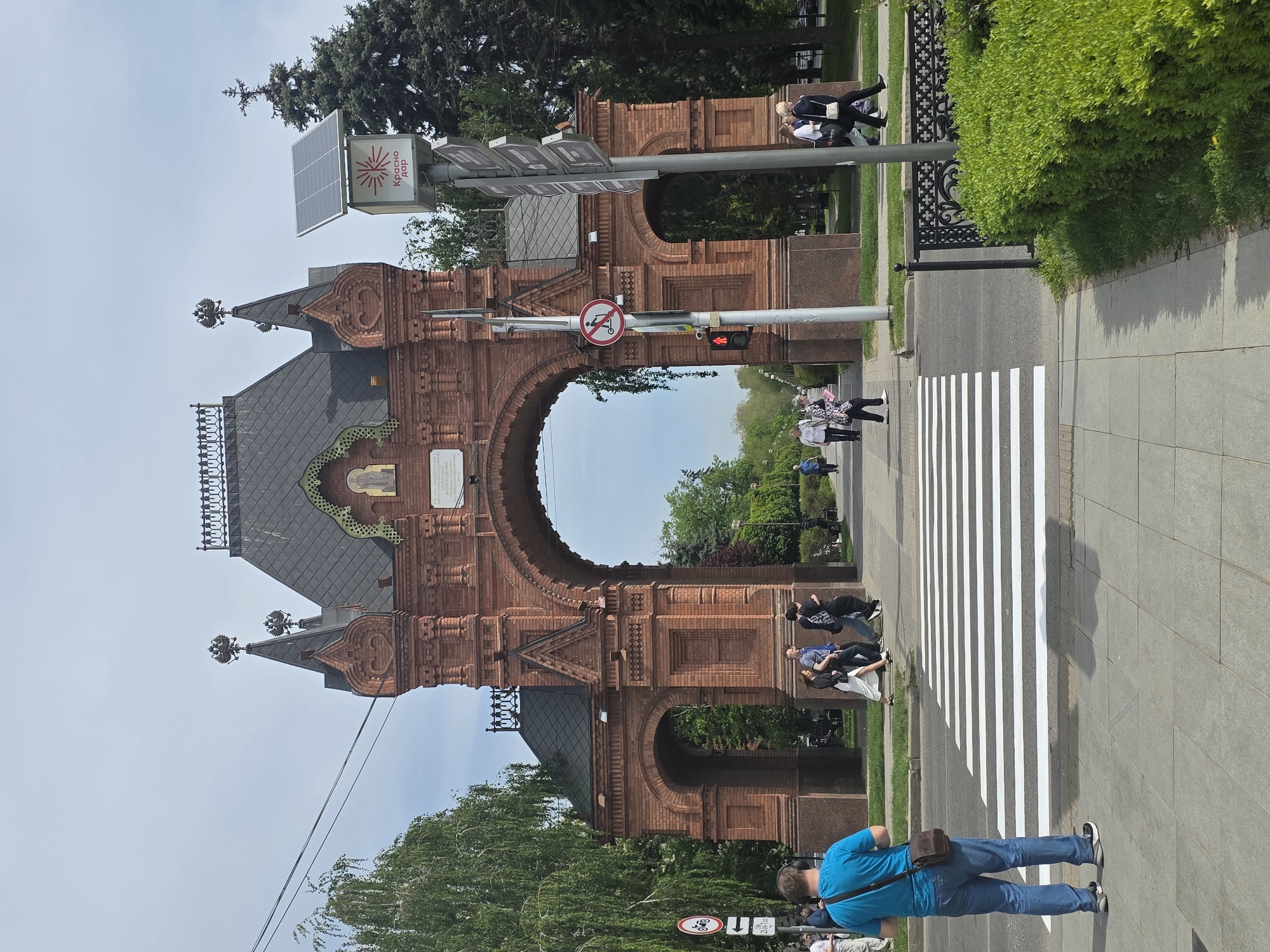 The image shows the Tsar Gate in the city of Krasnodar, Russia. The gate is constructed of red brick, with a large archway in the center. The roof is dark gray, and there are decorative elements such as double-headed eagles. A solar panel is attached to the right side of the gate. A road passes beneath the gate, and a crosswalk is visible with several people crossing. Other people are also visible on the sidewalks to either side. Street signs are present, including a "no motorcycle" sign and a sign reading "Krasnodar." The surrounding environment includes trees, bushes, and green spaces. A man wearing a blue shirt and jeans stands in the lower right corner, looking at his phone.