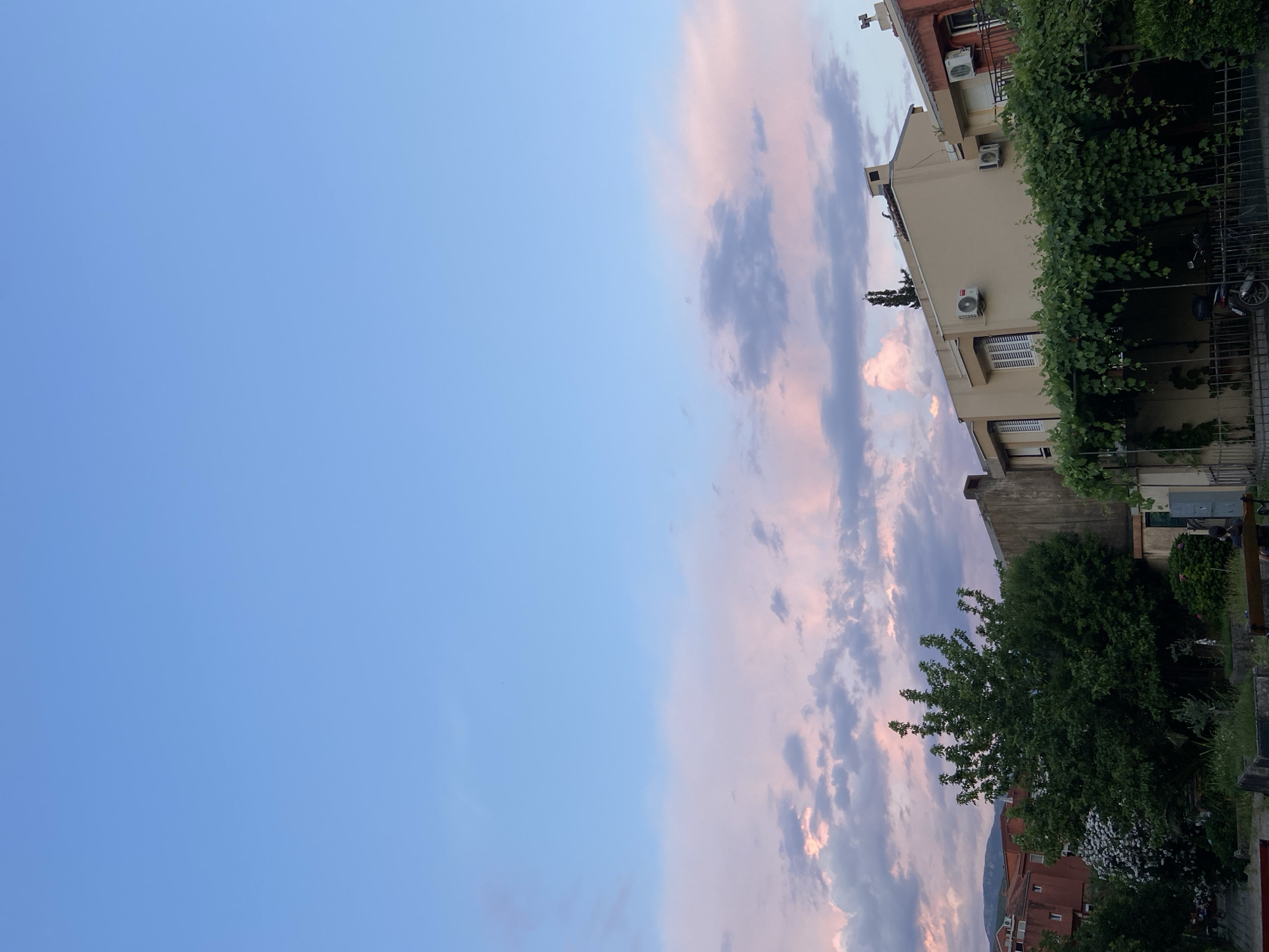 The image shows a view of buildings and trees under a sky with pink and purple clouds. The buildings appear to be residential homes, with several windows and air conditioning units visible. Green foliage covers a trellis in front of one of the buildings, and a tree stands tall to the left. In the foreground, there are hints of benches and a motorcycle. The sky is a gradient of light blue, transitioning to pastel shades of pink and purple, suggesting either sunrise or sunset. The overall impression is a serene, suburban setting.