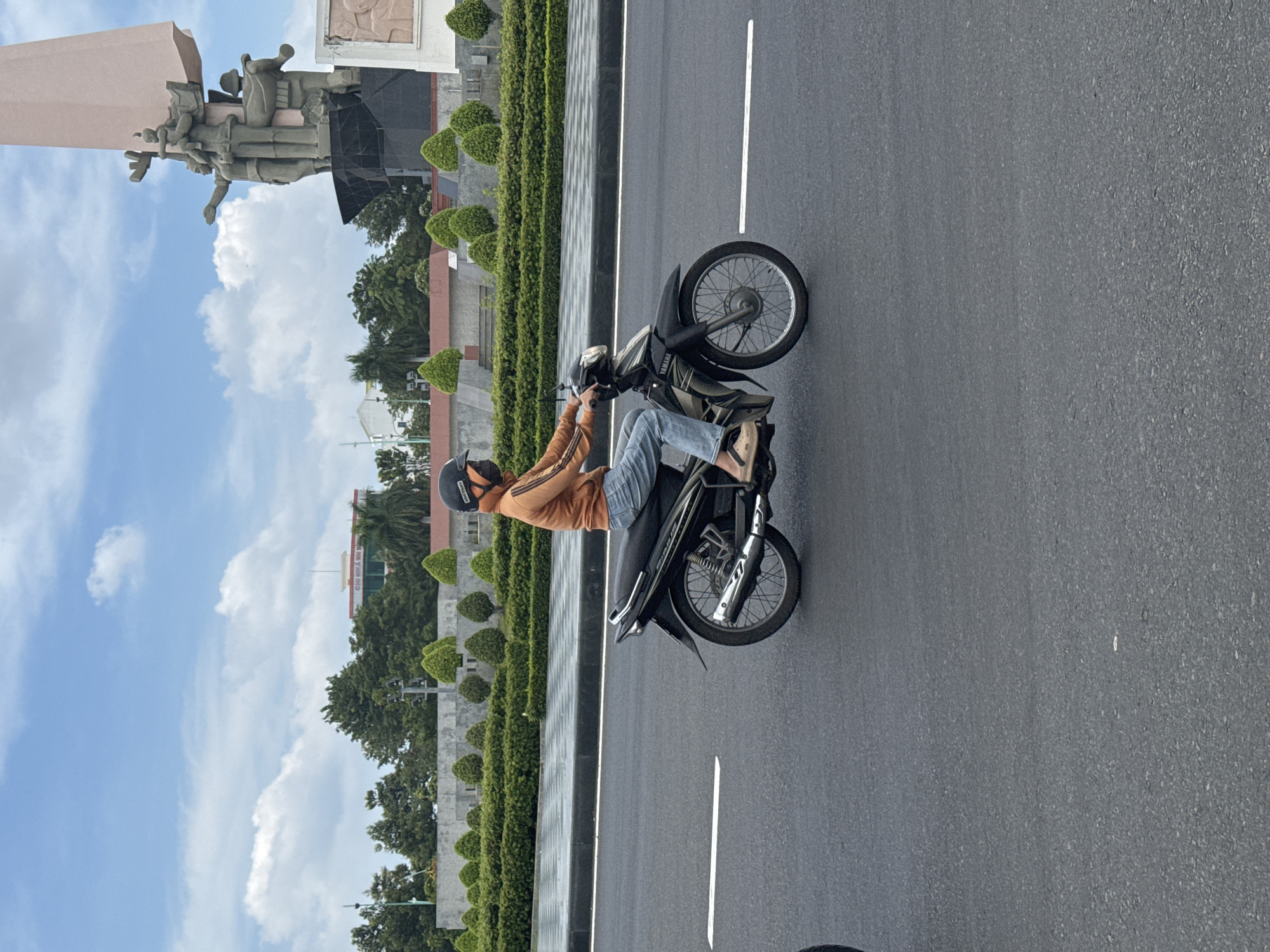 A person wearing an orange hooded sweatshirt, blue jeans, a black helmet, and a black face mask rides a black motorcycle on an asphalt road. The motorcycle has "YAMAHA" written on its side. In the background, there is a large monument with statues, lush green bushes, trees, a building with "CHÍNH HÃNG" written on it, and a cloudy blue sky. The scene is in Phan Thiết, Vietnam.