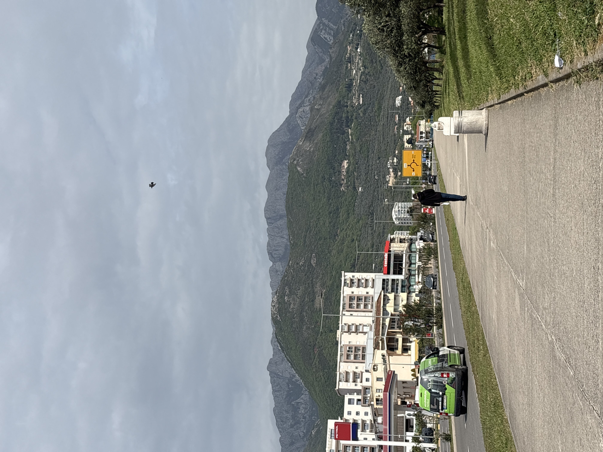 The image captures a vibrant street scene in a town, likely Bar in Montenegro, set against a dramatic backdrop of towering, rugged mountains. The perspective is taken from slightly above, looking down a wide, paved road that runs parallel to the mountains.
In the foreground, a concrete sidewalk with a narrow grassy verge extends along the right side of the road. A solitary person, dressed in a dark jacket and blue jeans, walks away from the viewer on the sidewalk. Further along the sidewalk, there are several concrete bollards or decorative posts.
To the left of the road, various buildings line the street. These include multi-story residential or commercial structures. A petrol station is visible, marked with a red "PETROL" sign. A green and white commercial van is parked near the petrol station, clearly displaying "VELA ALUMINIUM PVC FURNITURE" and a phone number on its side. Another building in the mid-distance has a red sign with "castell" visible.
A prominent yellow road sign stands by the road, featuring a roundabout symbol and providing directions to "CETINJE," "PETROVAC," and "BUDVA"—towns located in Montenegro.
The midground and background are dominated by an impressive range of mountains. Their lower slopes are densely covered in green vegetation, while the higher elevations reveal exposed, grey rock faces and peaks, stretching across the entire horizon. Various smaller buildings and structures are nestled within the green foothills of these mountains.
The sky is overcast and light grey, indicating a cloudy daytime. A small, dark bird is visible in flight in the upper left portion of the sky, adding a subtle touch of movement to the otherwise still scene.