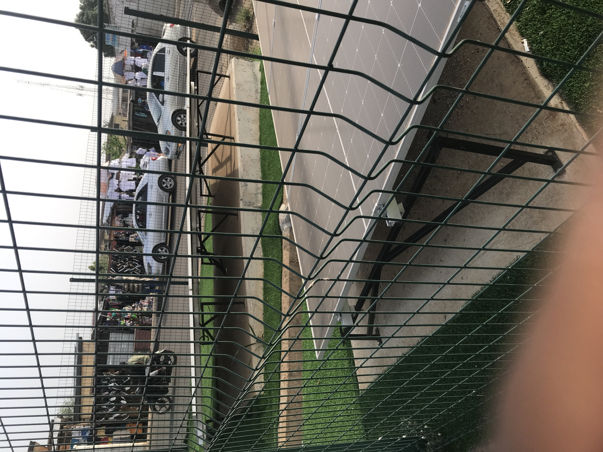 The image, rotated 90 degrees counter-clockwise, captures an outdoor scene viewed through a prominent green wire mesh fence in the foreground.
On the right side of the fence, several large, dark-grey solar panels are installed on metal frames over a concrete or paved surface. A narrow strip of artificial green grass runs along the base of the panels.
On the left side, beyond the fence, a busy street or open area is visible. Two white cars are parked or slowly moving. Around and between these cars, a group of people are gathered; many are dressed in white garments, possibly indicating school uniforms or traditional attire. Further into the background on this side, there are market stalls or small shops displaying various goods, suggesting a commercial area. A motorcycle is also visible parked closer to the foreground on the street side. In the distant background, a communication tower or similar structure stands against a bright, hazy sky.
The scene depicts everyday activity in what appears to be an urban or semi-urban setting, with modern infrastructure (solar panels) adjacent to a lively street and market. The time of day appears to be daytime, under bright, possibly overcast or hazy conditions, as there are no harsh shadows. No visible text is clearly discernible in the image.