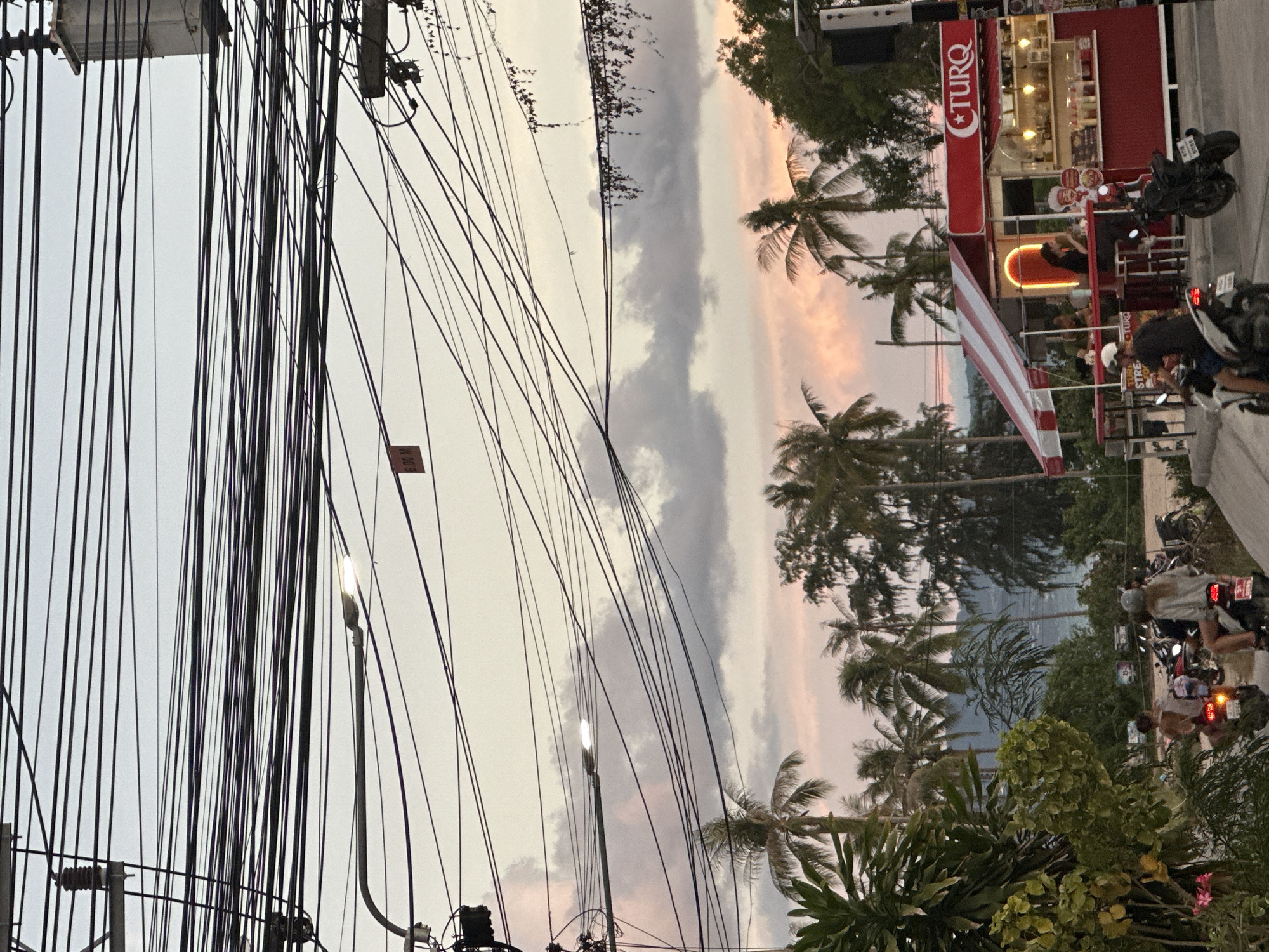 In the city of ตำบลเกาะพะงัน, Thailand, the image captures a street scene at dusk, with a prominent overhead tangle of electrical wires dominating the upper portion of the frame against a pale, cloudy sky tinged with sunset colors. Below the wires, palm trees and other tropical foliage line the street. In the mid-ground, a red and white striped food stall with a neon light accent operates, displaying the "CTURQ" logo. Several individuals are visible, with two people seated at the stall and a few others on motorcycles. One motorcycle in the foreground has two occupants, and another motorcycle is parked near the food stall. The overall impression is of a bustling, tropical urban environment transitioning into evening.