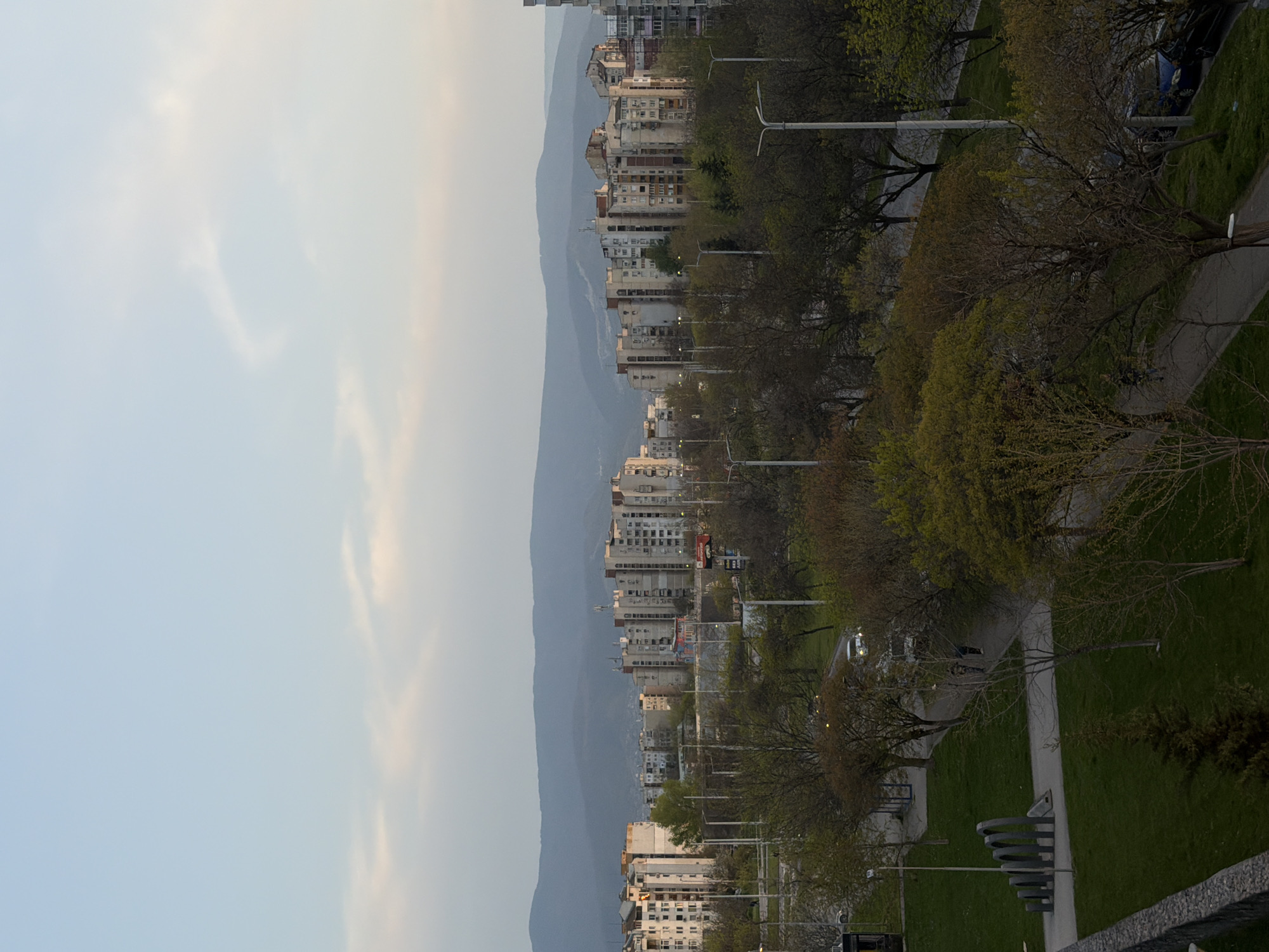 The image shows a cityscape in Plovdiv, Bulgaria. Buildings line the right side, with mountains in the background to the left. A row of trees and streetlights separates the buildings from a walkway and grassy area on the right. The sky occupies the left side of the image and is light blue.