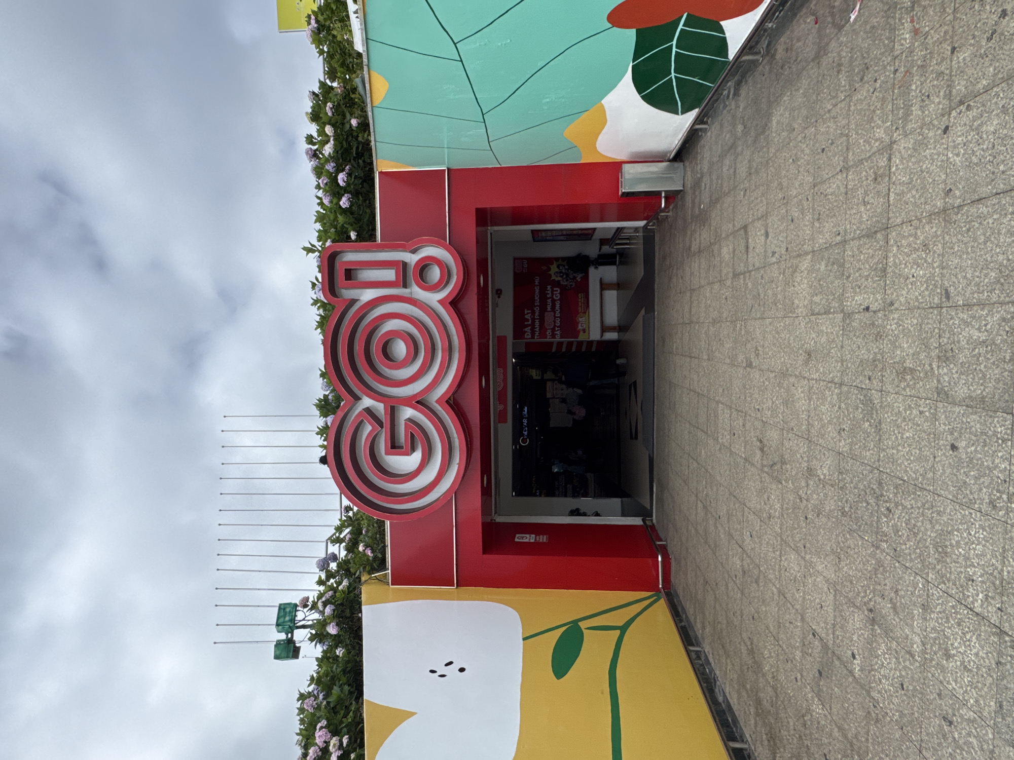 The image shows the entrance to a GO! supermarket in Da Lat, Vietnam. The entrance is marked by a large red "GO!" logo with a concentric circle design. The building is painted in bright colors, with a yellow wall on the left featuring a white flower illustration, and a teal wall on the right decorated with green leaves. Above the entrance, there are rows of flagpoles against a cloudy sky, and greenery with pink and purple flowers. The ground is paved with square, light-colored tiles. Inside the entrance, a sign indicates "ĐÀ LẠT THÀNH PHỐ SƯƠNG MÙ" (Da Lat, City of Fog) and "MUA SẮM GẦY GU DÙNG GU" (Shopping Near Gu). Several people are visible entering or exiting the supermarket.