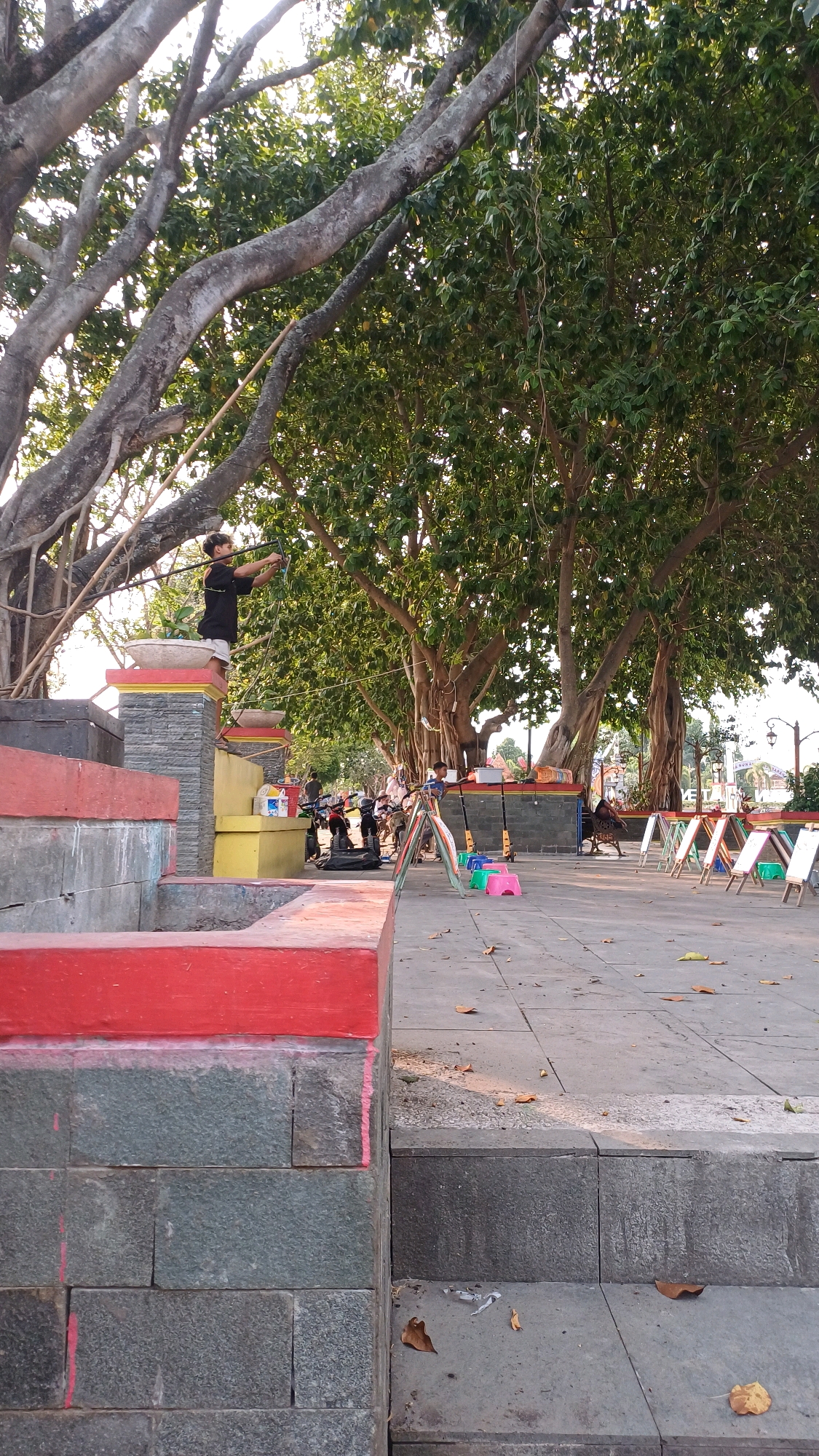A young person stands on a raised platform with a long stick in hand, possibly practicing archery, under the shade of large banyan trees in a public park in Klaten, Indonesia. The park features a paved area with several easels set up, suggesting an art or performance space, and a few children's swings. A few people are visible in the background, with motorcycles parked nearby, indicating it is a frequented public area. The foreground is dominated by tiered, stone and painted concrete structures, with red accents. The atmosphere appears to be relaxed and communal.