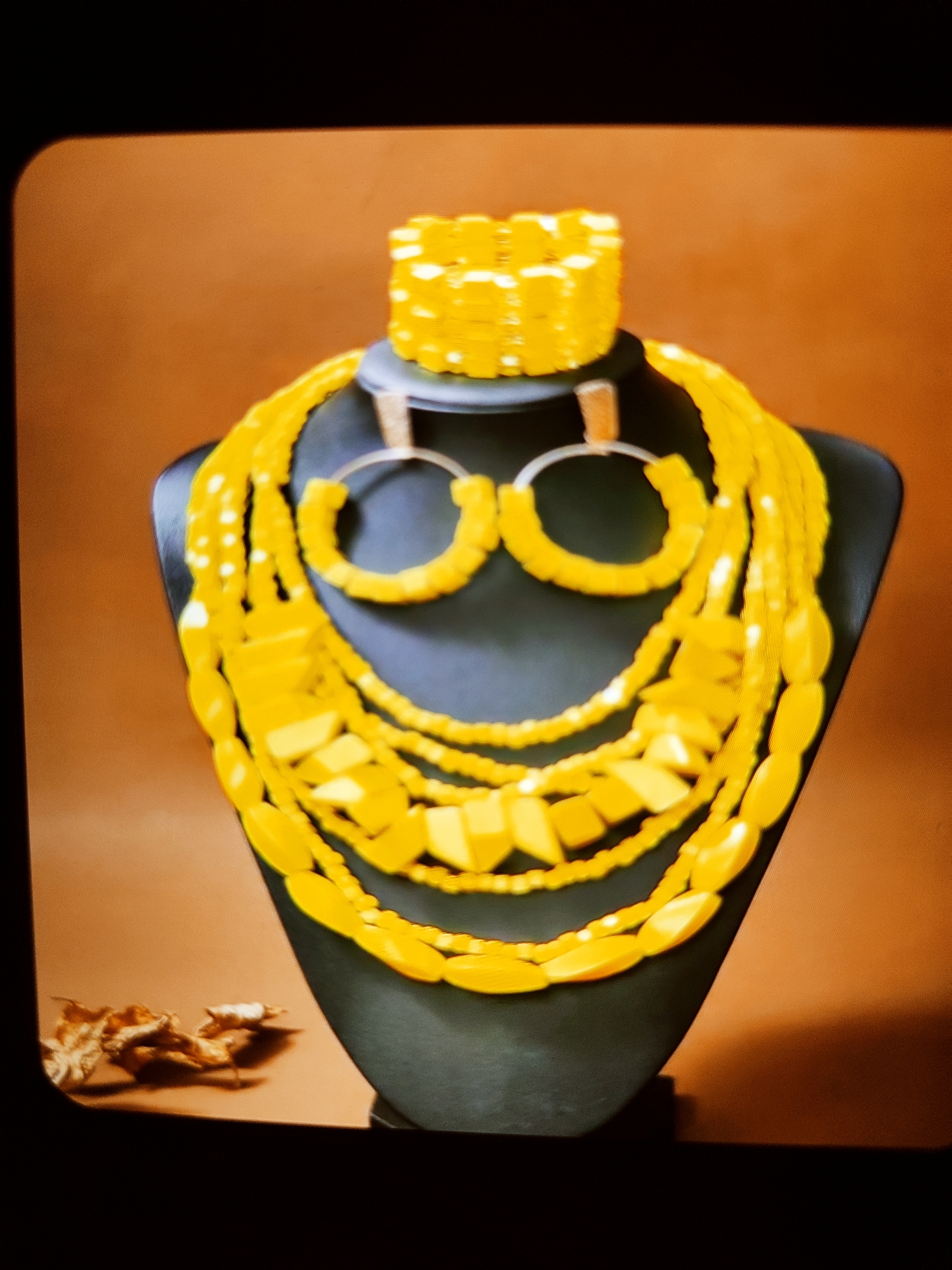 A yellow, multi-strand necklace set, including a bracelet and hoop earrings, is displayed on a black mannequin bust. The jewelry features beads of varying shapes and sizes, predominantly oval and rectangular. The background is a warm, light brown. In the lower left corner, two dried, brown leaves are visible on the surface. The scene is framed by a black border with rounded corners.