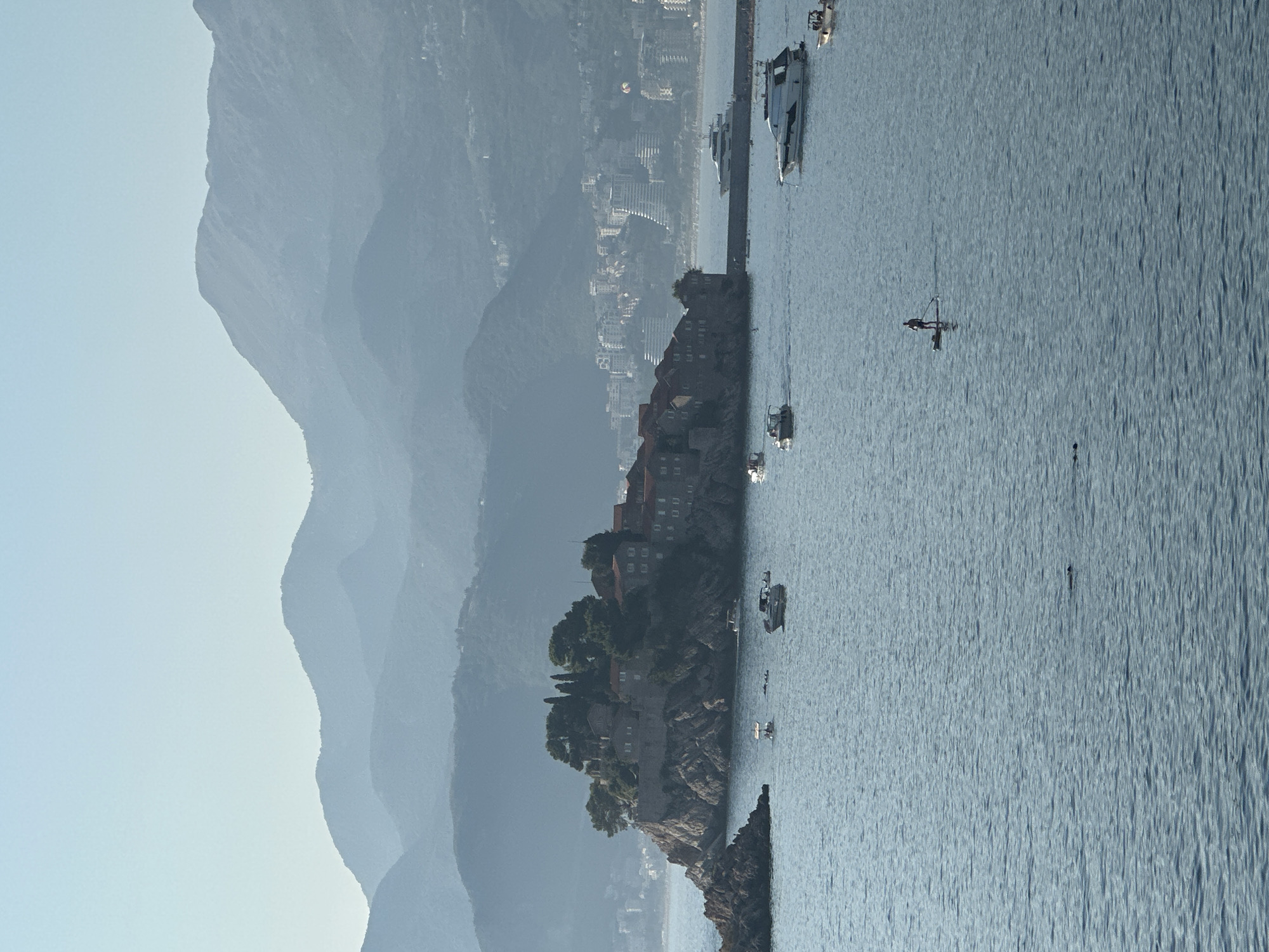 An aerial wide shot captures the island of Sveti Stefan, Montenegro, a picturesque peninsula dotted with stone buildings and lush greenery, connected to the mainland by a narrow causeway. Several boats, including a large white yacht, navigate the calm, blue waters surrounding the island. In the foreground, a lone individual stands on a paddleboard, silhouetted against the tranquil sea. The backdrop features a hazy mountain range, with a city skyline visible on the coast. The sky is a clear, pale blue, indicating a bright, sunny day.