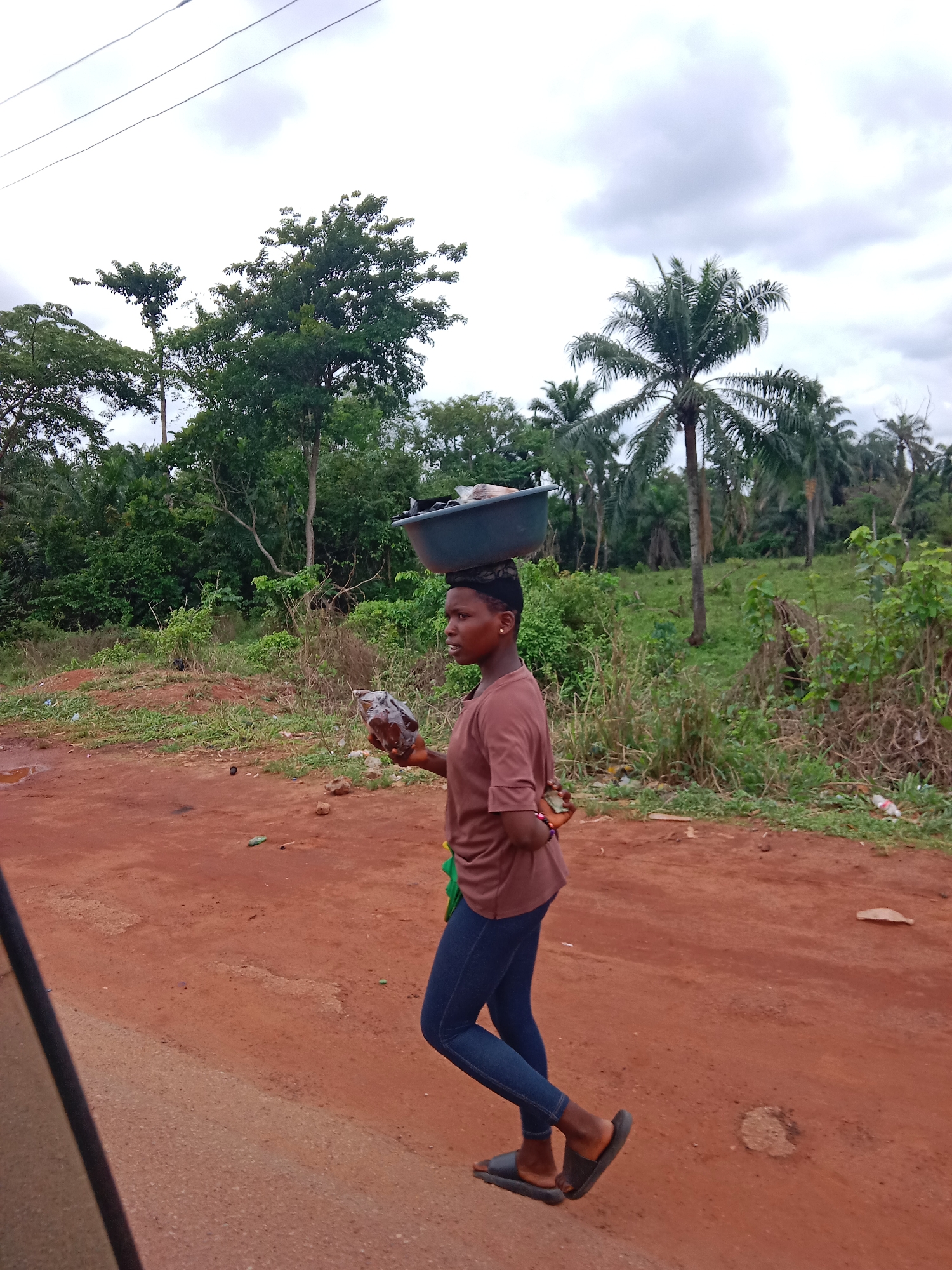 A young woman is walking on a dirt road. She is carrying a container on her head. She is wearing a brown shirt, blue jeans, and sandals. She is holding a plastic bag in her right hand. The road is dirt and the surrounding area is overgrown with vegetation. There are palm trees and other trees in the background. The sky is cloudy. There are power lines overhead. The city is Unknown, Unknown.