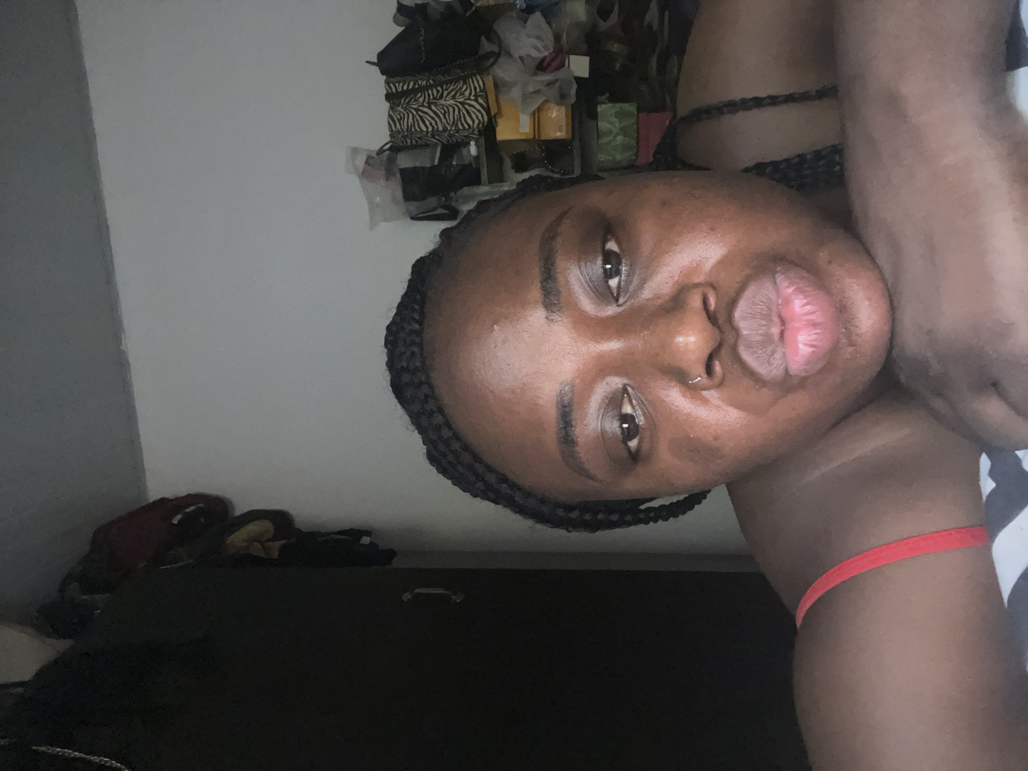 A close-up selfie features a person with dark skin and braided hair, positioned indoors. The image is rotated 90 degrees counter-clockwise. The subject looks directly at the camera, making a playful "duck face" or "kissing face" with puckered lips. A small silver nose ring is visible in their right nostril. Their skin shows a slight sheen, possibly from natural oils or direct lighting.
The person is wearing a red wristband on their right arm, and part of a black and white patterned top, possibly striped, is visible on their shoulder. The background is an indoor setting. On one side, there is a dark piece of furniture, possibly a dresser or cabinet, alongside a plain wall. On the other side, a cluttered shelving unit or storage area is visible, containing various items including several bags, one prominently featuring a black and white zebra print, as well as boxes and other unidentifiable objects. The lighting in the room appears somewhat dim, but the subject's face is well-lit. No text is visible in the image.