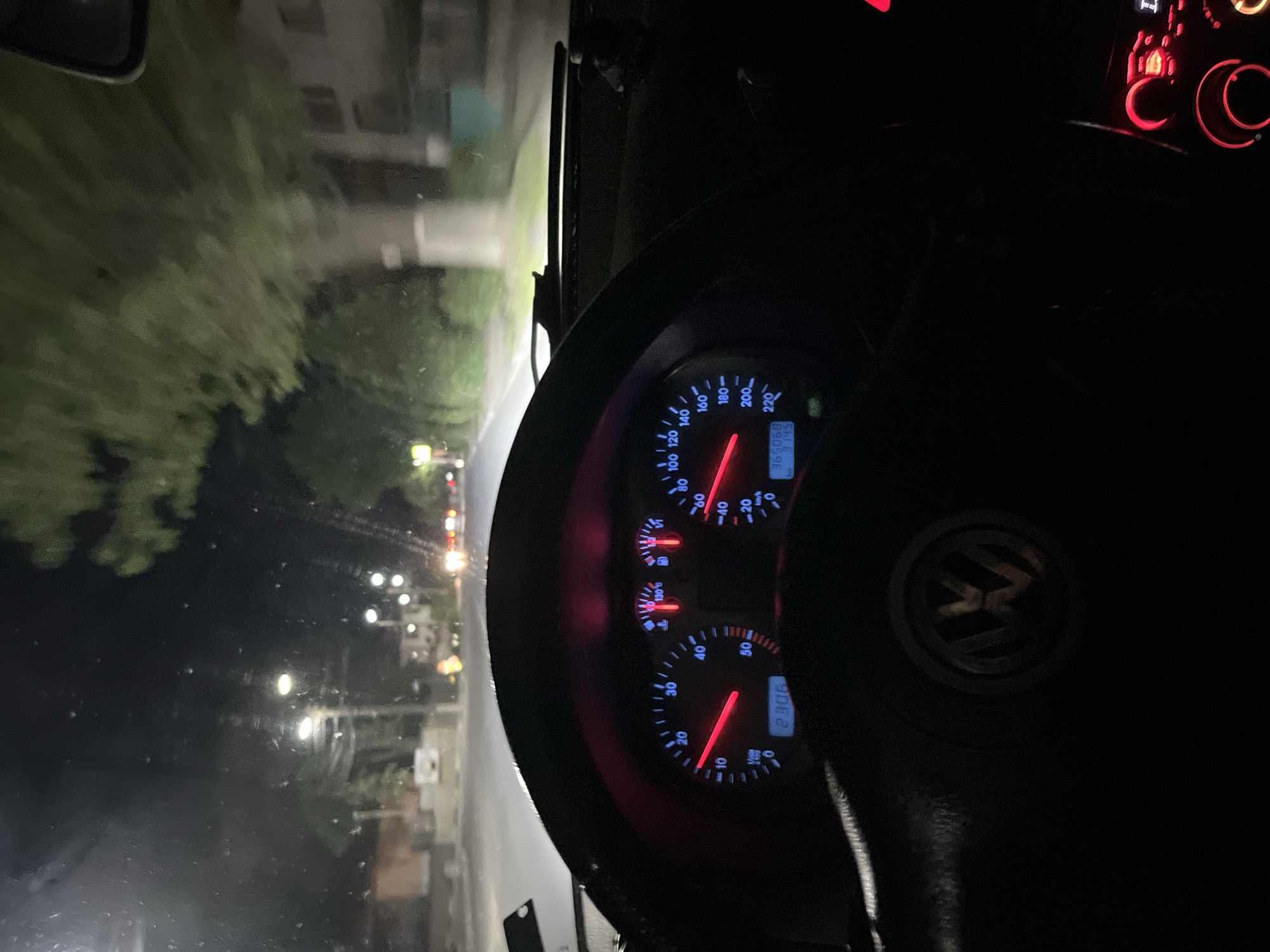 The image depicts the interior of a Volkswagen car, specifically the dashboard and steering wheel, at night. The speedometer reads approximately 75 km/h, and the tachometer reads about 2300 RPM. The odometer shows 365068 km and 3.145 km. The car is being driven on a street with streetlights and trees visible through the windshield. Based on the car model and the speedometer units, it is likely in Europe. The location is Anevo, Bulgaria.