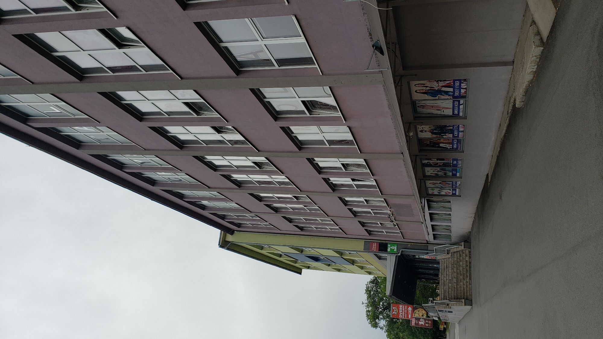 The image captures a two-story building with a purple facade and a yellow-green facade to its left, located on a street in Prešov, Slovakia. The purple building features numerous windows on both visible floors, with some appearing to be open. A series of posters, likely advertisements for fashion or a similar retail business, are displayed along the ground floor exterior wall of the purple building. To the left of the purple building, a gray staircase leads up to an entrance area with a dark awning, suggesting access to the buildings. Signs are visible near the entrance, one with "2+1 DVERE PODLAHY" and another with "PIZZA". The sky is overcast. The scene depicts an urban environment with commercial or institutional structures.