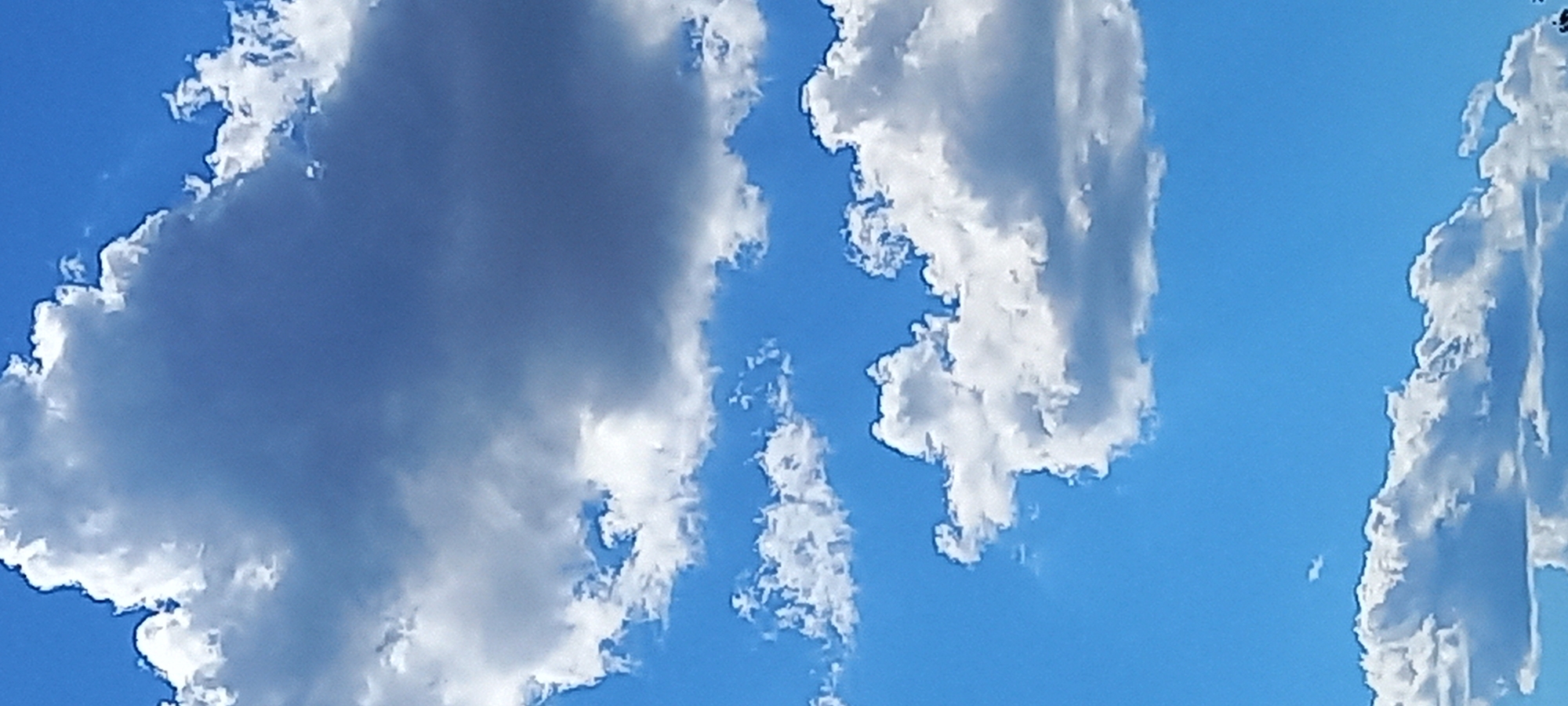 The image shows a bright blue sky with several white clouds of varying sizes and shapes. The clouds are fluffy and billowy, with some darker gray areas indicating shadows or thicker density. The scene is presumably located in Kryvyi Rih, Ukraine, based on the user's request. No people, subjects, or objects other than the sky and clouds are visible in the image. There are no actions or interactions depicted. The environment is a clear, sunny day.