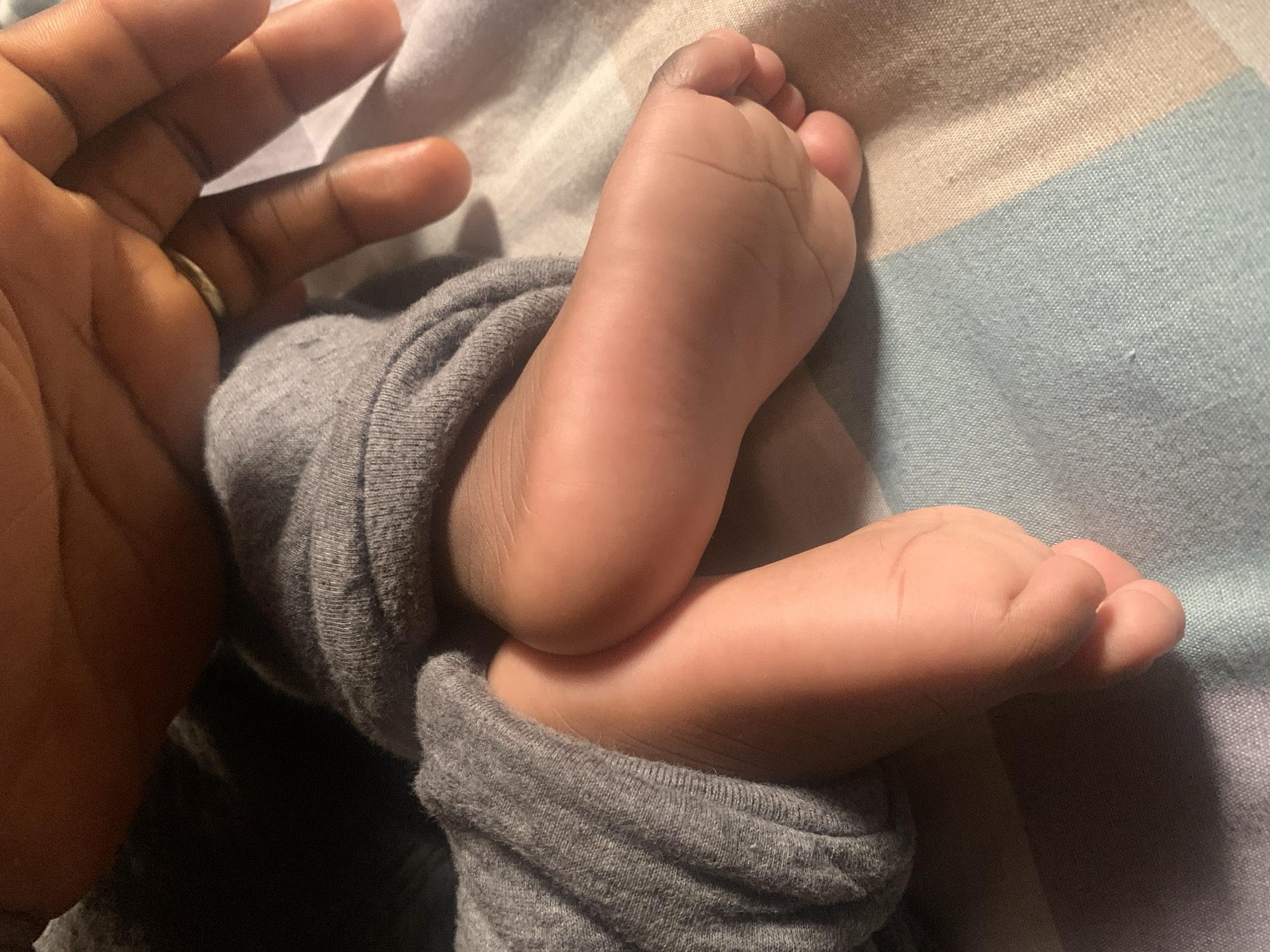 This close-up image portrays a tender moment between an adult and a baby in an indoor, cozy setting, possibly Jos, Nigeria, although no specific location cues are visible within the frame. On the left, a portion of an adult's hand is visible, featuring a dark skin tone and a gold-colored ring on the fourth finger. The hand appears to be gently resting near or cradling two small, plump baby feet, also with a dark skin tone, positioned towards the center and right of the frame.
The baby's feet are curled up, suggesting a state of rest or comfort. They are partially encased in soft, grey fabric, likely a baby's sleepsuit or pants, which is bunched around the ankles. The background consists of a striped fabric, likely bedding or a blanket, displaying soft horizontal stripes in shades of light blue, beige/cream, and off-white.
The lighting in the image is soft and diffused, creating a peaceful and intimate atmosphere, possibly indicating evening, early morning, or a dimly lit room. The overall impression is one of warmth, affection, and a loving connection between the adult and the baby. There is no visible text present in the image.