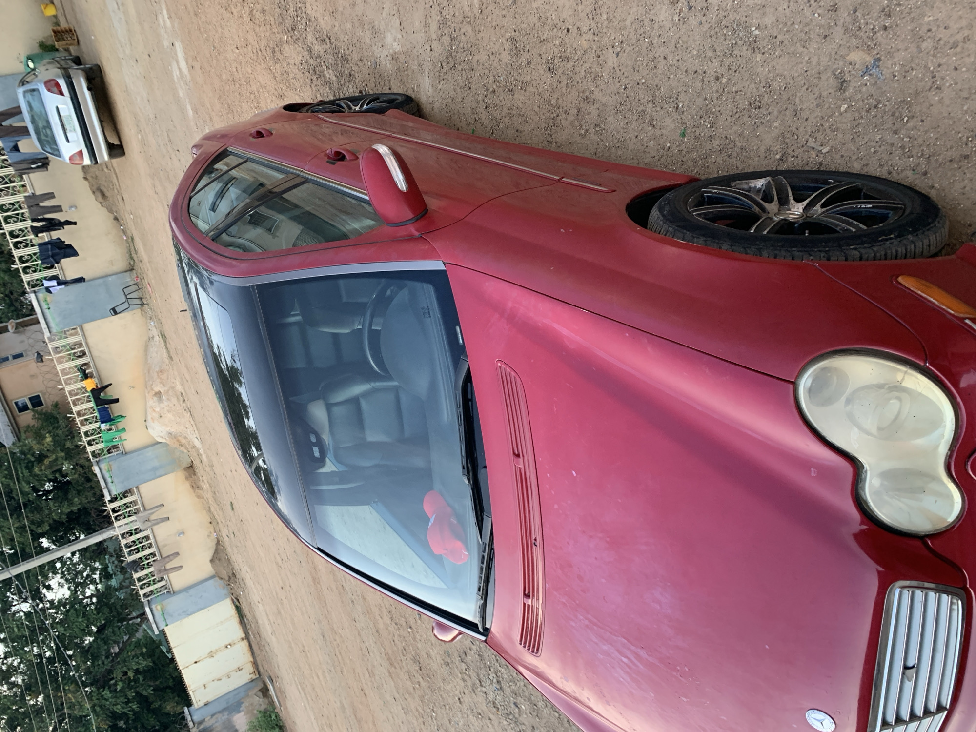 A red Mercedes-Benz coupe is parked on a dirt surface, possibly a roadside or parking area, in an urban environment. The car appears dusty. In the background, a second, silver/white vehicle is visible along with a building and what appear to be clothes hanging outside. The location is given as the city of New Stadium, Nigeria. No people are directly visible in the immediate foreground, although there might be individuals in the background near the building and second vehicle. A pair of red objects are sitting on the dashboard in the foreground.