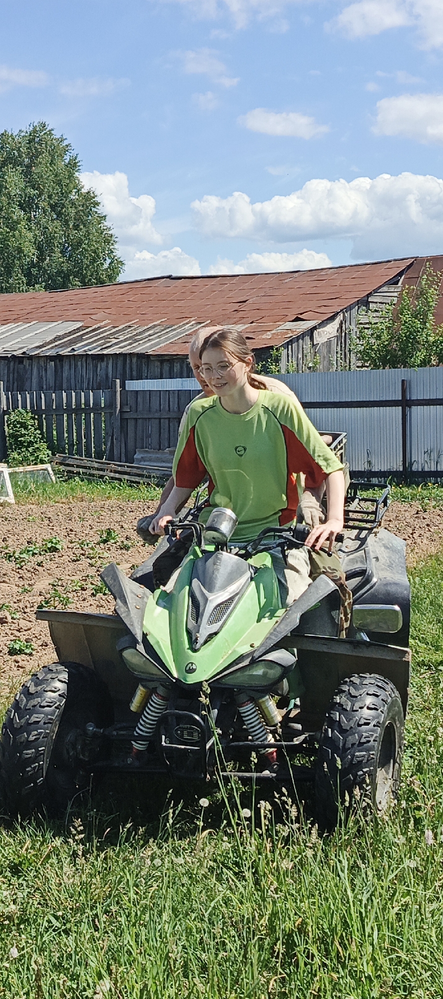 Two individuals are seated on a green and black all-terrain vehicle (ATV) outdoors in Vanki, Russia, under a clear blue sky with white clouds. The ATV is positioned on a patch of green grass with sparse weeds. To the left, a section of dark, tilled soil is visible.
The front rider is a young adult with light skin and dark hair, wearing round glasses, a bright green long-sleeve shirt with orange panels, and light-colored pants. Their left hand is on the left handlebar, and they are looking slightly to their right with a slight smile. The person behind them is an older adult, partially obscured, wearing a light-colored shirt. Their left hand, wearing a light-colored glove, rests on the right handlebar.
In the background, behind a weathered wooden fence, stands a dilapidated wooden building with a steeply pitched, rusty corrugated metal roof. To the right of the building, a light-colored corrugated metal fence is present. Lush green trees are visible to the far left and behind the fences. The scene is brightly illuminated by natural daylight.
