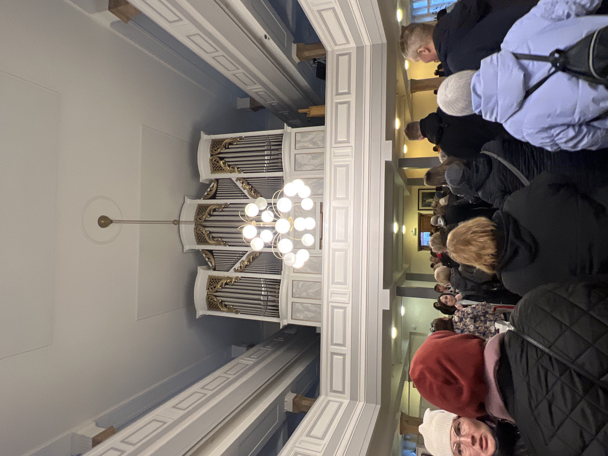 Inside a church-like building in Saint Petersburg, Russia, an audience is seated in pews, facing forward. A pipe organ, illuminated by a chandelier, stands above them. The architectural design of the space features tall ceilings, decorative molding, and a light color scheme.