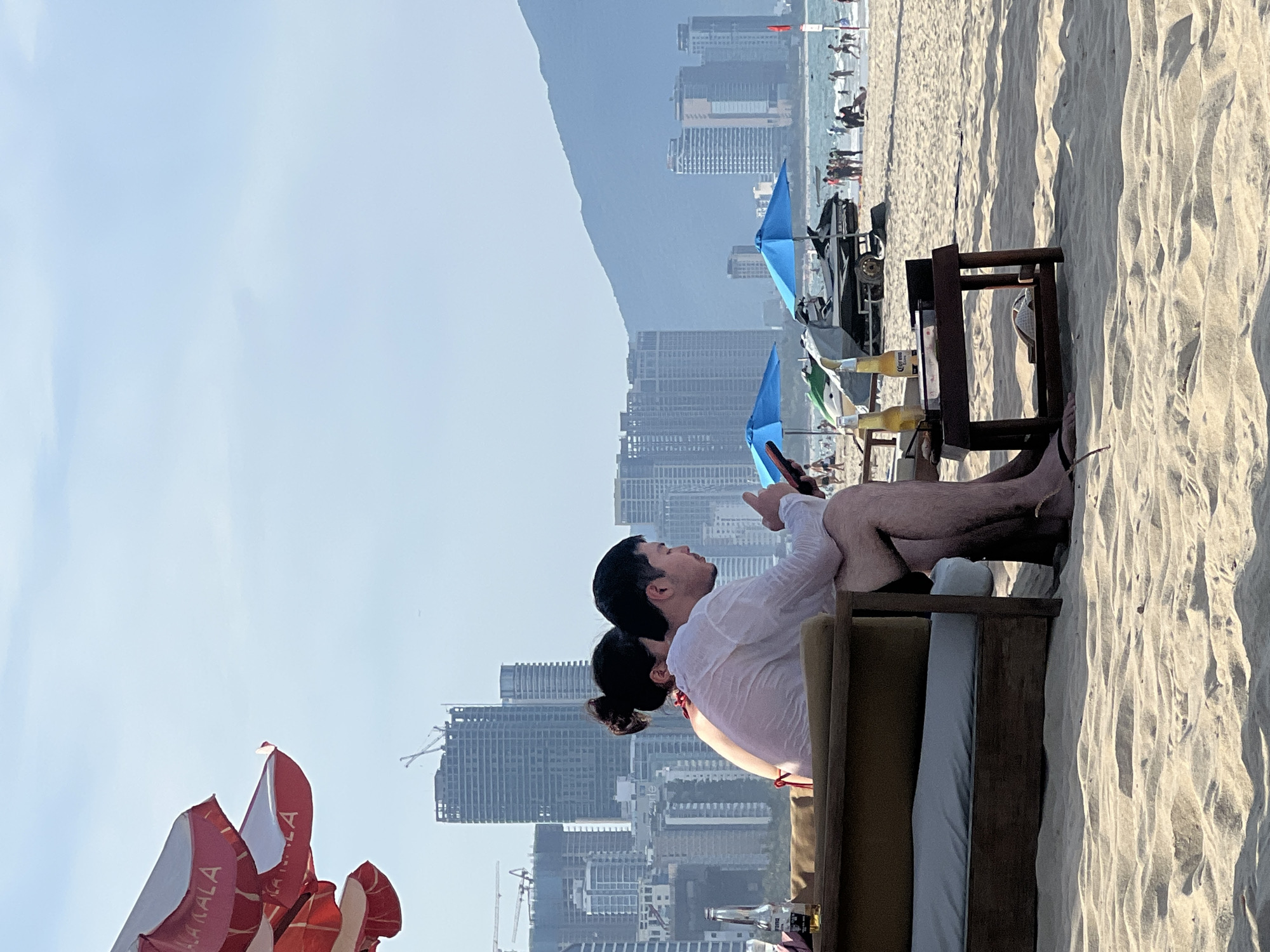 The image depicts a beach scene in Đà Nẵng, Vietnam, with the coastline and city skyline in the background. In the foreground, a couple is seated on a low, wooden couch on the sand, facing away from the viewer. The man, wearing a white shirt, is looking down at his phone. The woman, with her hair tied up, is leaning against him, wearing a red bikini top. Beside them, on a small wooden table, are two bottles of beer and a pair of flip-flops.
The beach is dotted with other beachgoers, umbrellas, and jet skis. In the distance, the modern architecture of Đà Nẵng's skyscrapers rises against a hazy sky, with construction cranes visible on some buildings, indicating ongoing development. A mountain range is visible in the far distance, adding to the natural landscape that contrasts with the urban sprawl. The overall atmosphere suggests a leisurely day at the beach with urban views.