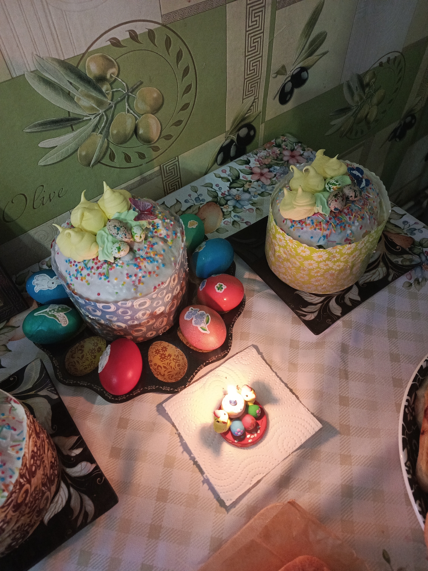 The image presents a still life composition of Easter traditions, likely taken indoors in a home in Chaikovsky, Russia, given the cultural context. Two elaborately decorated Easter cakes, known as kulich, are prominently displayed. The cakes are frosted with white icing and adorned with colorful sprinkles, meringue toppings, and decorative Easter eggs.
One kulich sits on a decorative black tray surrounded by colorful Easter eggs, some adorned with floral and geometric patterns. Another kulich rests on a black tray with a floral pattern. The background features a wall with a decorative pattern of olive branches and olives, and floral wallpaper is behind the cakes.
In the foreground, a small candle holder in the shape of Easter eggs and chicks sits on a white paper napkin. The lighting is soft, creating a warm and festive atmosphere. The items are arranged on a white and gray checkered tablecloth. The presence of kulich and decorated eggs suggests the scene captures a celebration of Easter.