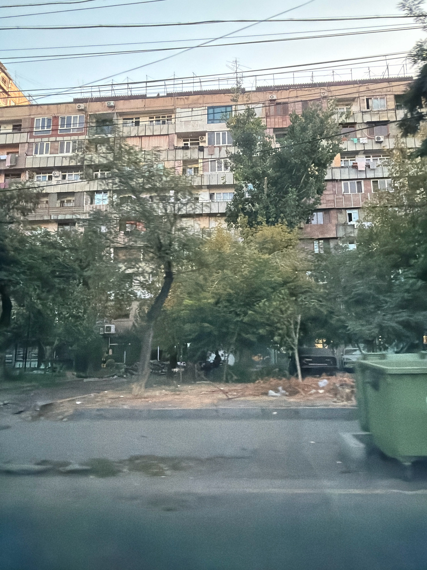 The image depicts an aging, Brutalist-style apartment building in Yerevan, Armenia. The building, characterized by its repetitive concrete facade and numerous balconies, appears to be of Soviet-era architecture. Clutter is visible on many of the balconies, including drying laundry, air conditioning units, and various household items. Several trees with green foliage are positioned in front of the building, partially obscuring it. In the foreground, a street is visible, with a green dumpster to the right. The sky above is overcast with visible power lines. The overall scene suggests an urban environment in Yerevan, likely in a residential district.