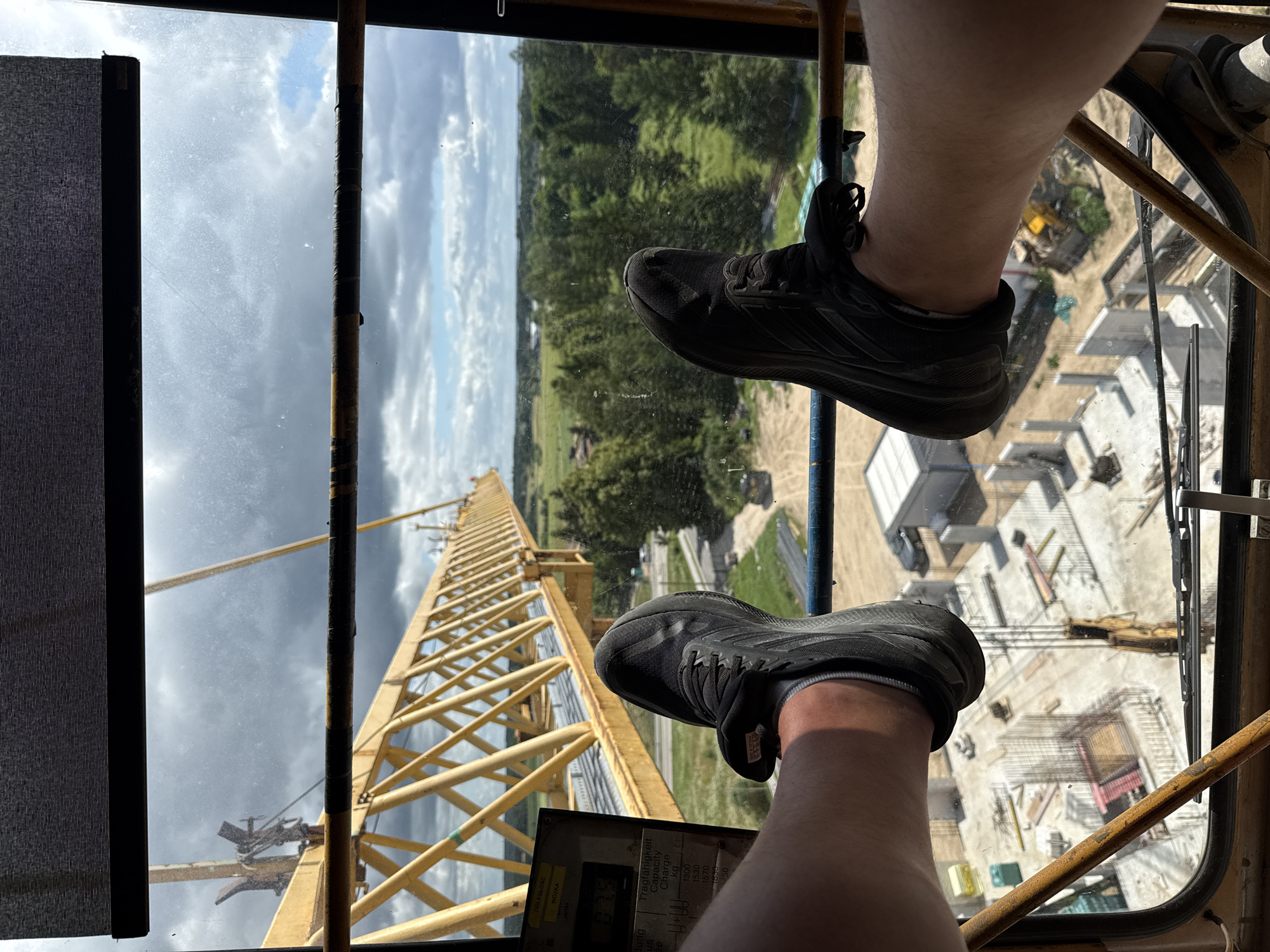 The image is a first-person perspective shot from inside a construction crane cabin, looking out at a construction site. Two legs, clad in black athletic shoes and white socks, are propped up on the cabin's railing, obscuring much of the foreground. The crane's yellow boom extends horizontally across the frame. Below, a construction site is visible, featuring concrete foundations, rebar cages, and what appear to be building structures in various stages of completion. Construction equipment, including a yellow excavator, is present. The background shows a rural landscape with trees and fields under a cloudy sky. A control panel with Polish text, including "Dźwigniołe" (lever) and "UDŹWIG" (load) is partially visible on the left side of the cabin. The context suggests the image was taken in Mikołajki, Poland, likely during a work break on a construction project.