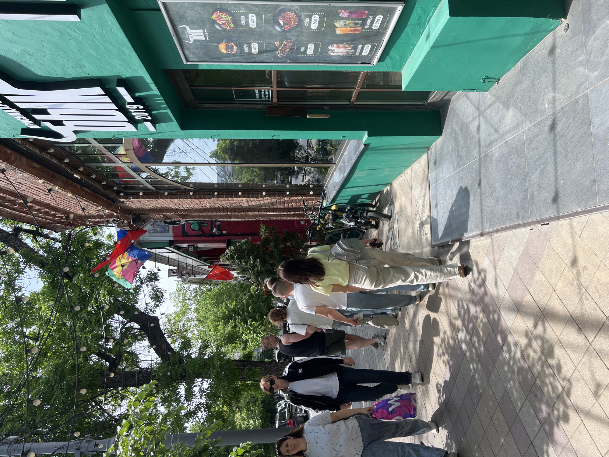 Here is a description of the image:
The image depicts a scene from a city street, likely in Krasnodar, Russia, as indicated by the Cyrillic text on a restaurant's menu board. The restaurant has a green facade with a menu board displaying items like burgers, salads, ribs, and steaks with their prices in rubles.
A line of people is standing on the sidewalk in front of the restaurant. There appear to be at least six people in the queue, with varying styles of dress. In the background, there's a tree with fairy lights and several colorful flags. There are also parked bicycles near the building.