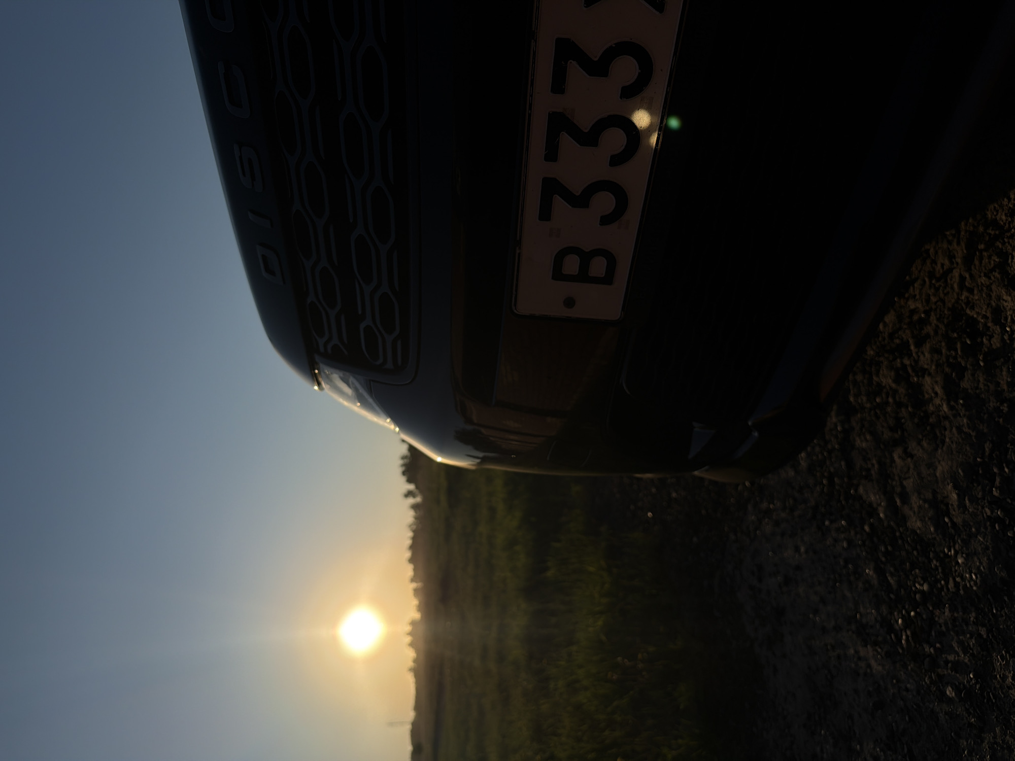 A dark-colored vehicle, likely a Land Rover Discovery due to the visible "DISC" lettering on its body, is positioned in the foreground of a rural landscape. The image is oriented vertically, showing the front or side of the car, which features a distinctive hexagonal patterned grille. A Russian license plate bearing the sequence "B333" is clearly visible on the vehicle, with a small bright reflection near the numbers.
The setting is an open field at sunset. A bright, radiant sun is low on the horizon, casting a warm glow and creating a noticeable lens flare. The sky above is a clear blue, transitioning to lighter hues towards the sun. The sun's light illuminates a vast green field, beyond which a line of trees is silhouetted against the horizon. The ground adjacent to the vehicle appears to be a dark, textured dirt or gravel road. The scene suggests a calm evening in a rural area near Krasnoyarsk, Russia. No specific activity is taking place; the vehicle appears stationary.