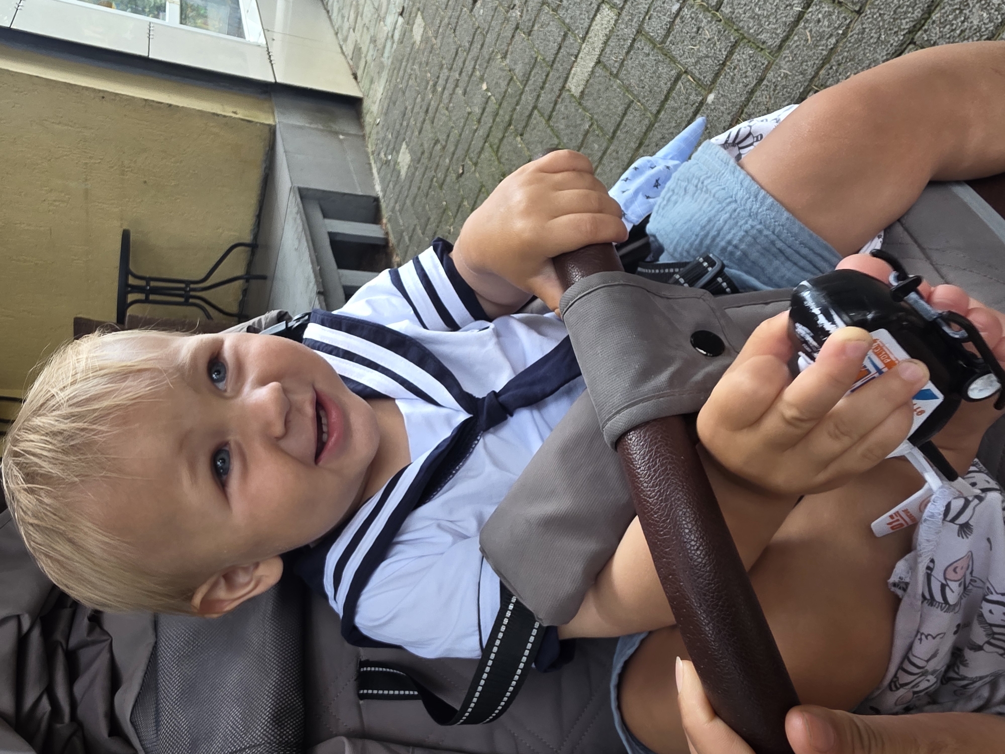 A baby with blonde hair and blue eyes is sitting in a stroller. The baby is wearing a sailor-style outfit with navy blue stripes on a white shirt. The baby is holding a small black toy with red and white details, possibly a toy car or vehicle, in their right hand. The baby is looking at the camera with a happy expression, with their mouth open as if speaking or laughing.
The stroller appears to be made of a brown leather-like material for the handle, and gray fabric. The baby is wearing light blue shorts and a patterned cloth with black and white zebra designs is visible underneath. The background shows a paved surface made of interlocking stones, suggesting an outdoor setting. Behind the stroller, a portion of a yellow building with a window is visible, and to the right, a stone wall and some steps leading upwards.
The lighting suggests it is daytime, likely overcast or shaded, as there are no harsh shadows. The overall mood is cheerful and captures a candid moment of a baby enjoying playtime outdoors. No visible text is clearly legible on the toy or clothing. Given the context, this scene is located in Sochi, Russia.