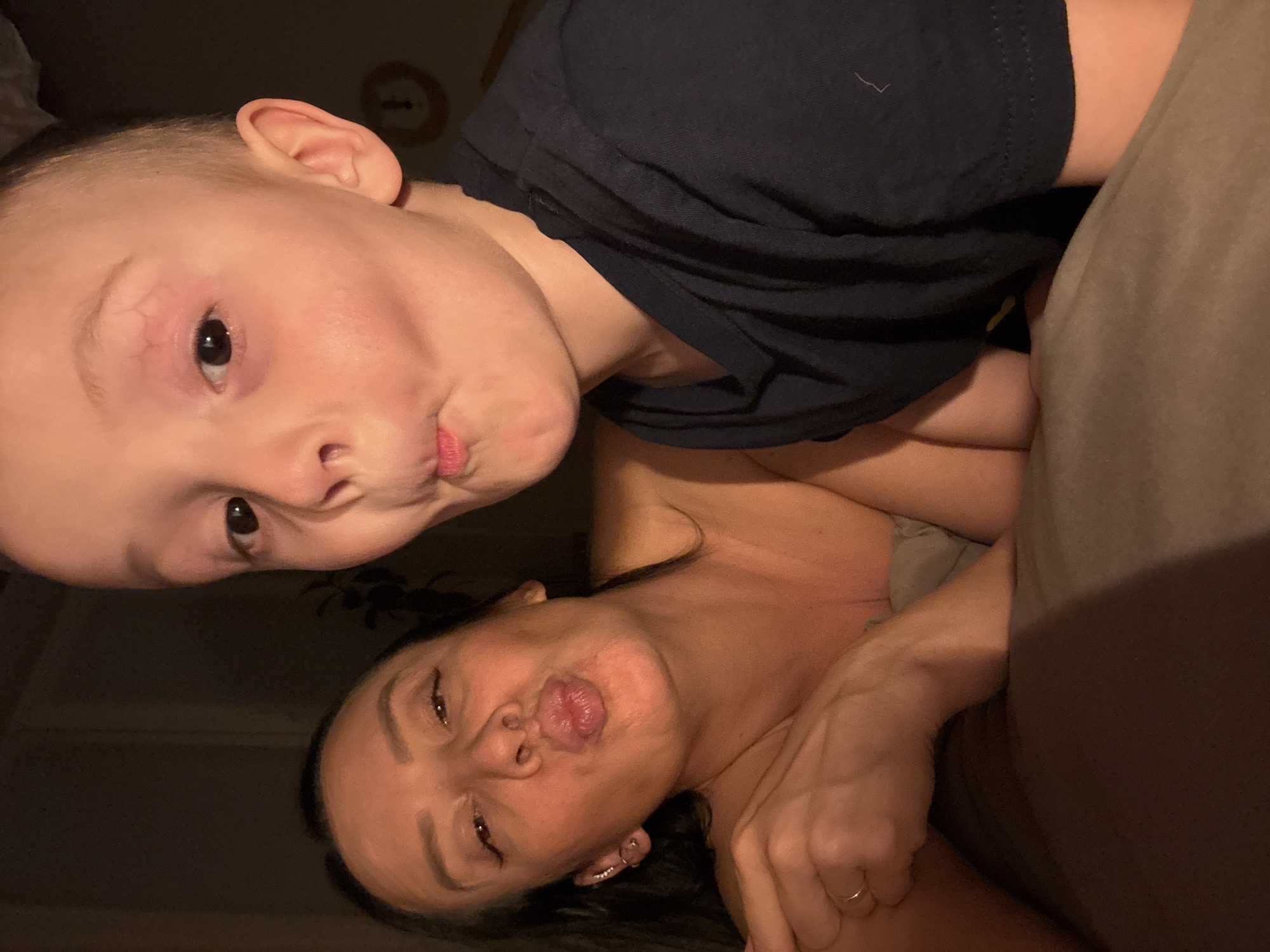 The image features two individuals, a young child and an adult woman, posing for a close-up, rotated selfie. Both are positioned horizontally and appear to be lying down, looking directly at the camera with playful, pouting or "kissing" facial expressions.
The child, with fair skin and light-colored hair, is in the upper portion of the frame. They are wearing a dark blue t-shirt. Notable details on the child include clearly visible veins around their left eye and a pronounced pout with slightly parted lips.
The adult woman is positioned below the child. She has dark hair and is also making a similar pouting face. A ring is visible on her right hand, and an ear piercing is visible on her left ear. Her bare shoulder or a light-colored top is visible.
The scene appears to be indoors, likely in a dimly lit room, suggesting an evening or nighttime setting. A brown or tan-colored blanket or pillow is partially visible on the right side of the frame, contributing to the impression of a relaxed, domestic environment. The overall mood is affectionate and playful. No specific objects or text are visible to indicate the Chaikovsky, Russia location.