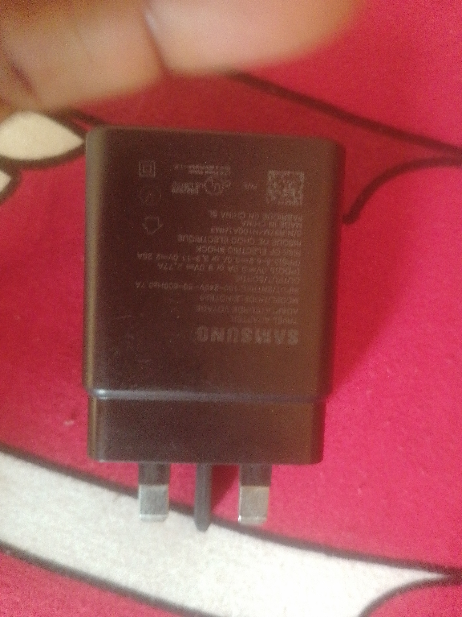 The image is a close-up, slightly blurry, overhead shot of a black Samsung travel adapter with UK-style plug pins. The adapter is resting on a red and white patterned surface, likely a fabric. Markings on the adapter include "SAMSUNG", "TRAVEL ADAPTER", "MODEL: EHOT820", "INPUT/ENTREE: 100-240V~50-60Hz0.7A", "OUTPUT/SORTIE: 5V=== 2A or 9V=== 1.67A or 3.3-5.9V=== 3A or 3.3-11V=== 2.77A", "RISQUE DE CHOC ELECTRIQUE", "MADE IN CHINA", "FABRIQUE EN CHINE", and other symbols and serial numbers. The location is specified as Danbazau, Nigeria.