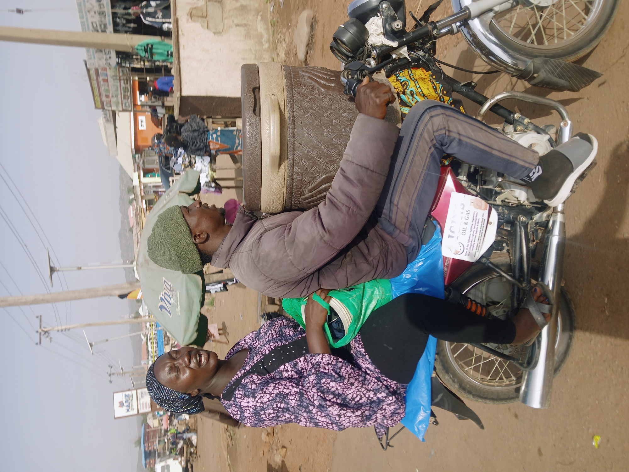 The image captures a lively street scene in Rafin Par, Nigeria, under bright, clear daylight. Two individuals, a man and a woman, are depicted on a motorcycle, which appears to be used for transportation and carrying goods.
The man, who is driving, wears a dark brown padded jacket, dark striped trousers, and dark slip-on shoes. A green beanie covers his head as he grasps the motorcycle handlebars. Behind him, a substantial, two-toned brown and beige cooler or insulated box is strapped to the back of the motorcycle.
The woman, riding as a passenger, is dressed in a black top adorned with a pink and white pattern, black leggings, and sandals. Her head is covered with a dark, polka-dotted headscarf. She holds a green shopping bag and has a large blue plastic bag draped over her leg. Her face expresses a wide, joyful smile or laugh, suggesting a cheerful mood.
The setting is a busy street, likely in a town or village, characterized by dusty, unpaved ground. In the background, there are various low-rise buildings, some appearing to be shops, and power poles with overhead lines. Faintly visible signage includes "Viva" on an umbrella and "HARZ" on a building. A notable detail is a white advertisement attached to the motorcycle's front fender, clearly displaying the text "101 OIL & GAS" with smaller print detailing services like "Sales, Drilling, Mining, etc." The scene depicts typical daily activity and transport in the area.