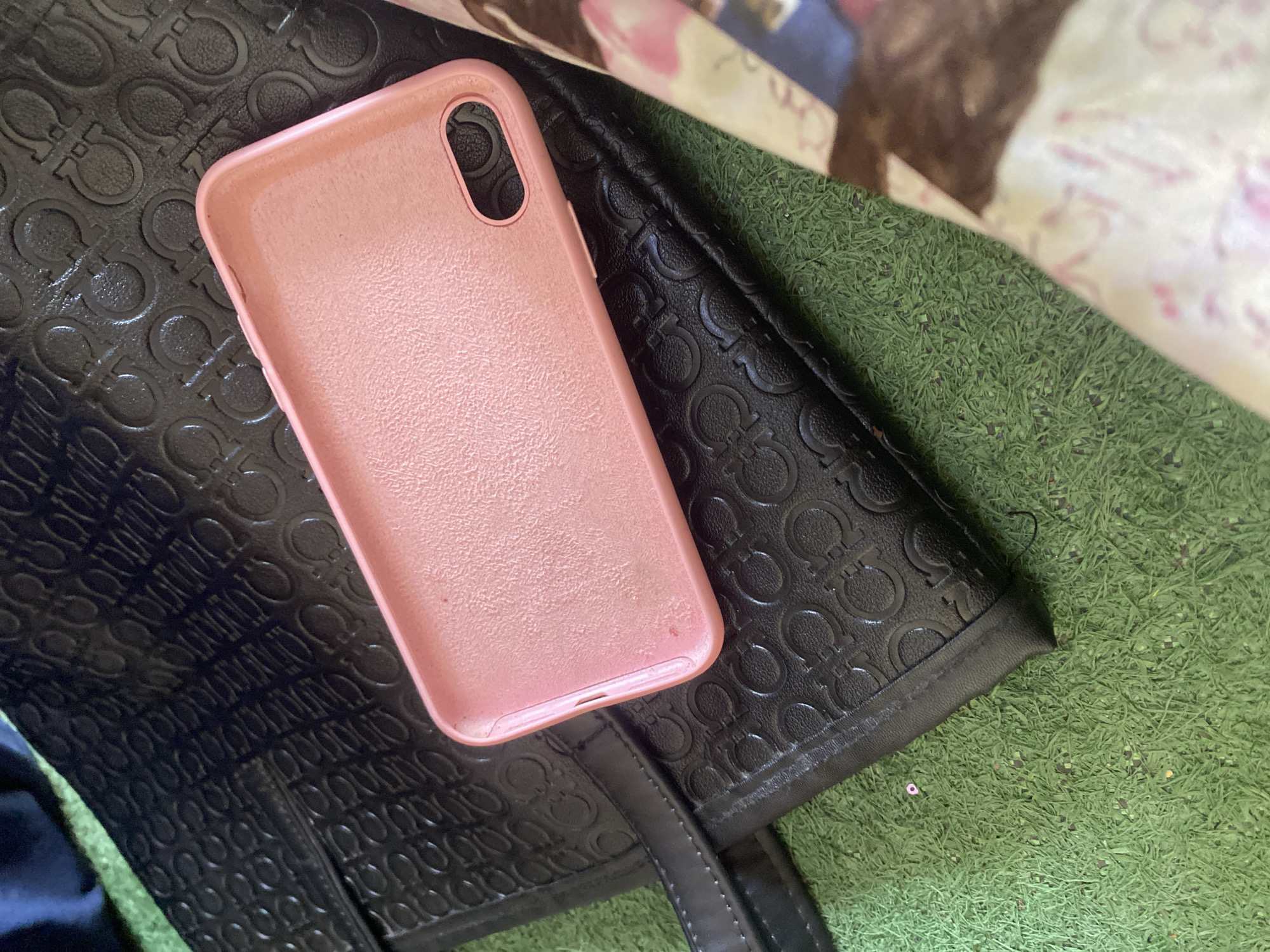 A pink phone case lies on top of a black handbag with an embossed pattern. The handbag is placed on a green, artificial turf surface. To the right of the handbag is a piece of patterned fabric. The scene appears to be outdoors, possibly in a yard or recreational area. There are no people visible in the image. The location in Jauro Yinu, Nigeria, is not discernible from the visual information provided.