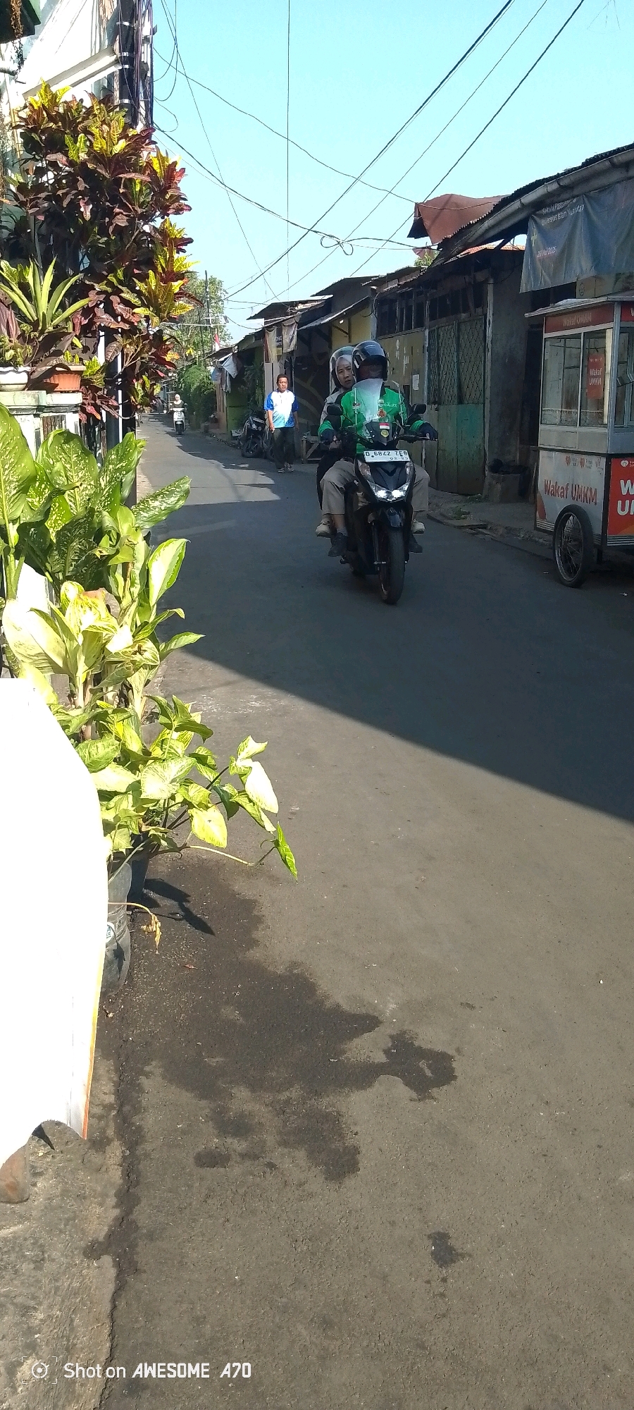 The image shows a street scene in Bandung City, Indonesia. In the foreground, there are potted plants with large green and yellow leaves. The street is paved and appears to be a residential area with buildings lining both sides. Several people and vehicles are visible. A motorcycle with two riders, both wearing helmets and the driver wearing a green jacket with branding, is the most prominent subject. The driver of the motorcycle is a man and the passenger is a woman. Further down the street, another person wearing a blue and white shirt is standing. There is also a food cart visible on the right side of the street with the words "WAKAF UMKM" printed on it. The sky is clear and blue, indicating a sunny day. The scene depicts a typical day in a bustling Indonesian city.