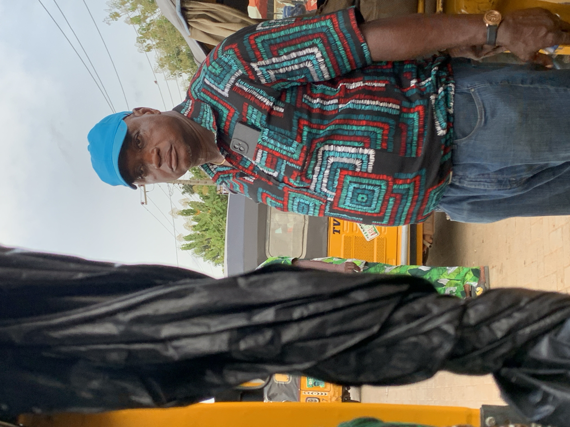 A medium shot depicts an older man wearing a blue cap, a patterned shirt, and jeans, standing outdoors. The man has a cell phone attached to his shirt. A vehicle, yellow with a license plate, is visible behind the man. The image also contains an object wrapped in black plastic in the foreground. The location is Ikorodu, Nigeria, as indicated by the vehicle's license plate.