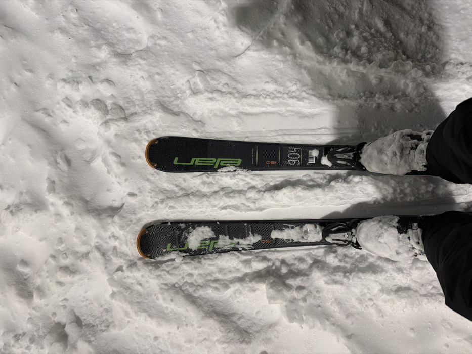 An overhead, first-person perspective captures an individual standing in deep, textured white snow in Makarovo, Russia. The person is wearing black ski pants and black ski boots, both of which are significantly covered with accumulated snow. Two black Elan brand skis are attached to the boots; these skis feature green branding and orange accents at their tips and tails. The right ski displays the markings "904" and "150", while the left ski shows "150". The bindings on both skis are visible and partially obscured by snow. The surrounding snow surface appears undisturbed, indicating a static position.