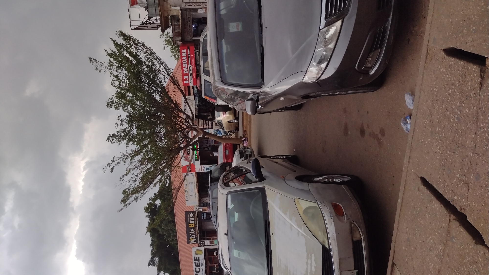 The scene captures a street in Jos, Nigeria, characterized by parked vehicles along the roadside. A silver car is parked in the foreground to the lower part of the image, another car is parked closer to the viewer, to the upper part of the image, a dark gray or black color. The vehicles line up alongside buildings housing local businesses, whose signs are legible in the image, the sky is overcast. Text on signage includes brands such as "B.B DANGANA COSMETICS," "White House," "TOT IDS SOLUTION," "CEE Fashion World." The overall atmosphere suggests a typical urban street scene in a developing area. The image appears to have been taken at around 15:30 based on the display of an electronic time sign.