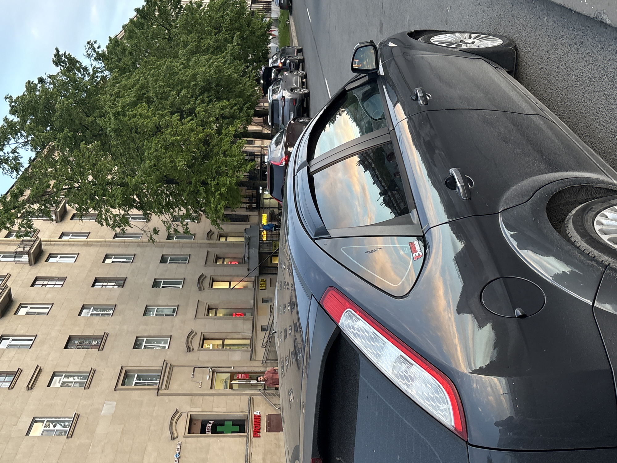 A street scene captures a dark grey or black car parked prominently in the foreground, angled to the right. The car's metallic surface reflects a sky with soft, warm clouds, indicating late afternoon or early evening. The rear taillight, fuel cap, and a "R3 Racing" sticker on the rear window are visible. The side windows also reflect the sky and parts of the adjacent buildings.
To the left, a multi-story building with numerous rectangular windows forms a strong vertical element, appearing light brown or beige. A large, lush green tree stands in front of and above the building, its leaves contrasting with the structured architecture.
Further down the street, a line of other parked cars is visible. Near an entrance of the building, a person in a reddish-brown top can be seen. Several commercial signs are affixed to the building's facade. One prominent sign features the text "АПТЕКА" (Pharmacy) next to a green cross symbol. Above it, another sign appears to read "ПРОМЫ". The overall impression is an urban environment, likely a city street, captured on a clear day.