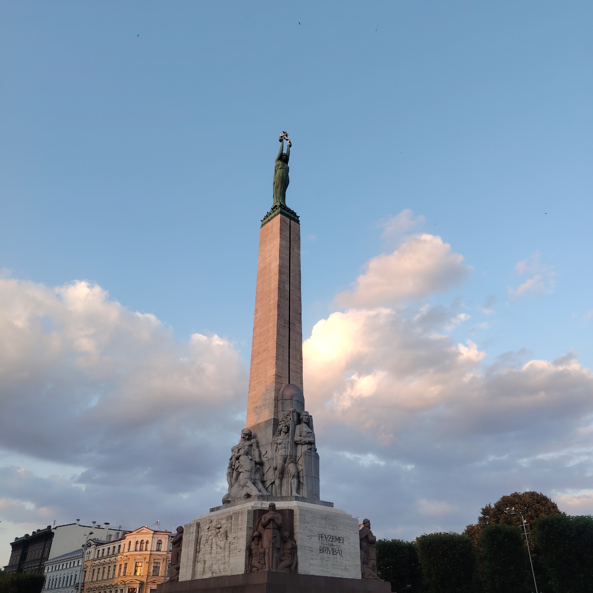 The image shows the Freedom Monument in Riga, Latvia, situated in the center of the city. The monument, a tall obelisk topped with a statue of a woman holding three stars, stands against a backdrop of a blue sky with scattered clouds. The monument itself is made of stone and features intricate carvings of figures at its base. In the foreground, there are buildings with traditional architecture and lush green trees. Several birds are visible in the sky. The lighting suggests it might be late afternoon or early evening, as the sun casts a warm glow on the monument and buildings.