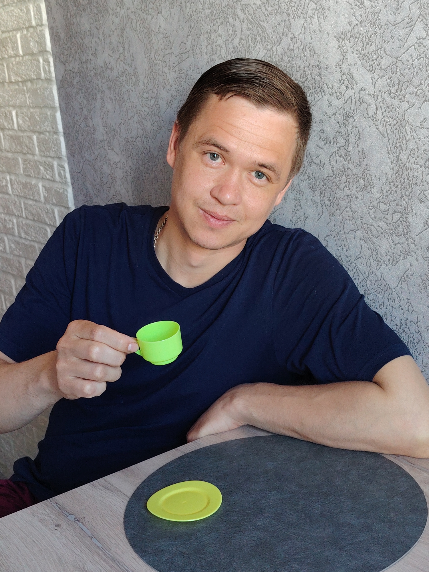 A fair-skinned man with short brown hair and blue eyes sits at a table indoors, holding a small, bright green toy teacup in his left hand. A small, yellow toy saucer rests on a dark grey circular placemat on the table. The man wears a dark blue t-shirt and a thin silver chain around his neck. The walls behind him are textured; one section is a light grey brick pattern, while the other is a light grey with a mottled texture. The table is light-colored with a wood grain pattern. The location is Chaikovsky, Russia.