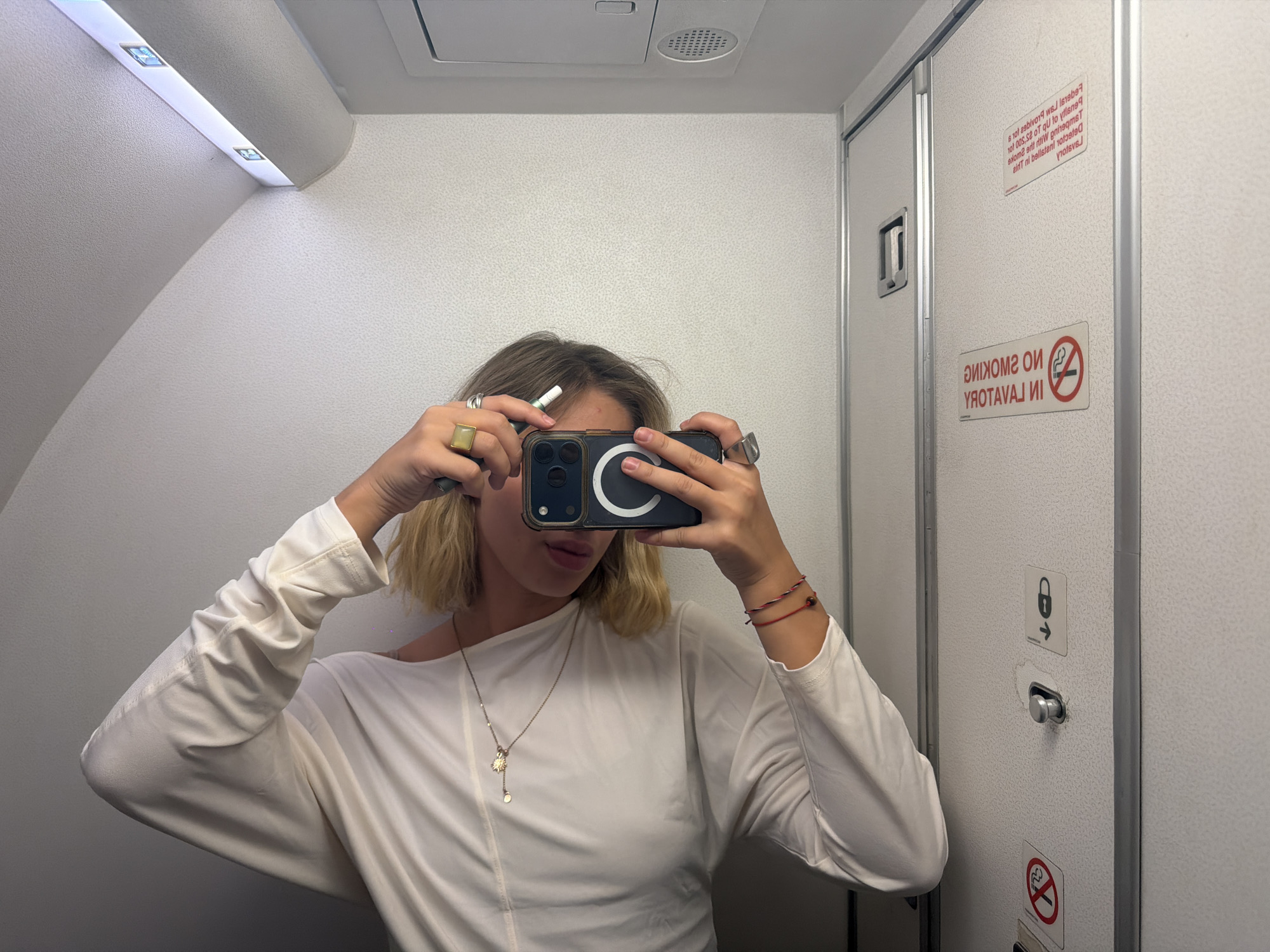 A young Caucasian woman with blonde hair tied into a ponytail takes a selfie in a mirror, holding a smartphone and a white pen. She is wearing a long-sleeved white off-the-shoulder shirt, a pendant necklace, and rings on her fingers, and red string bracelets on her wrist. The background appears to be a small lavatory, possibly on an airplane, with a white door on the right featuring a "No Smoking" sign. There are also lights visible on the ceiling and a ventilation grate. The image was taken in Cheremushka, Kazakhstan.