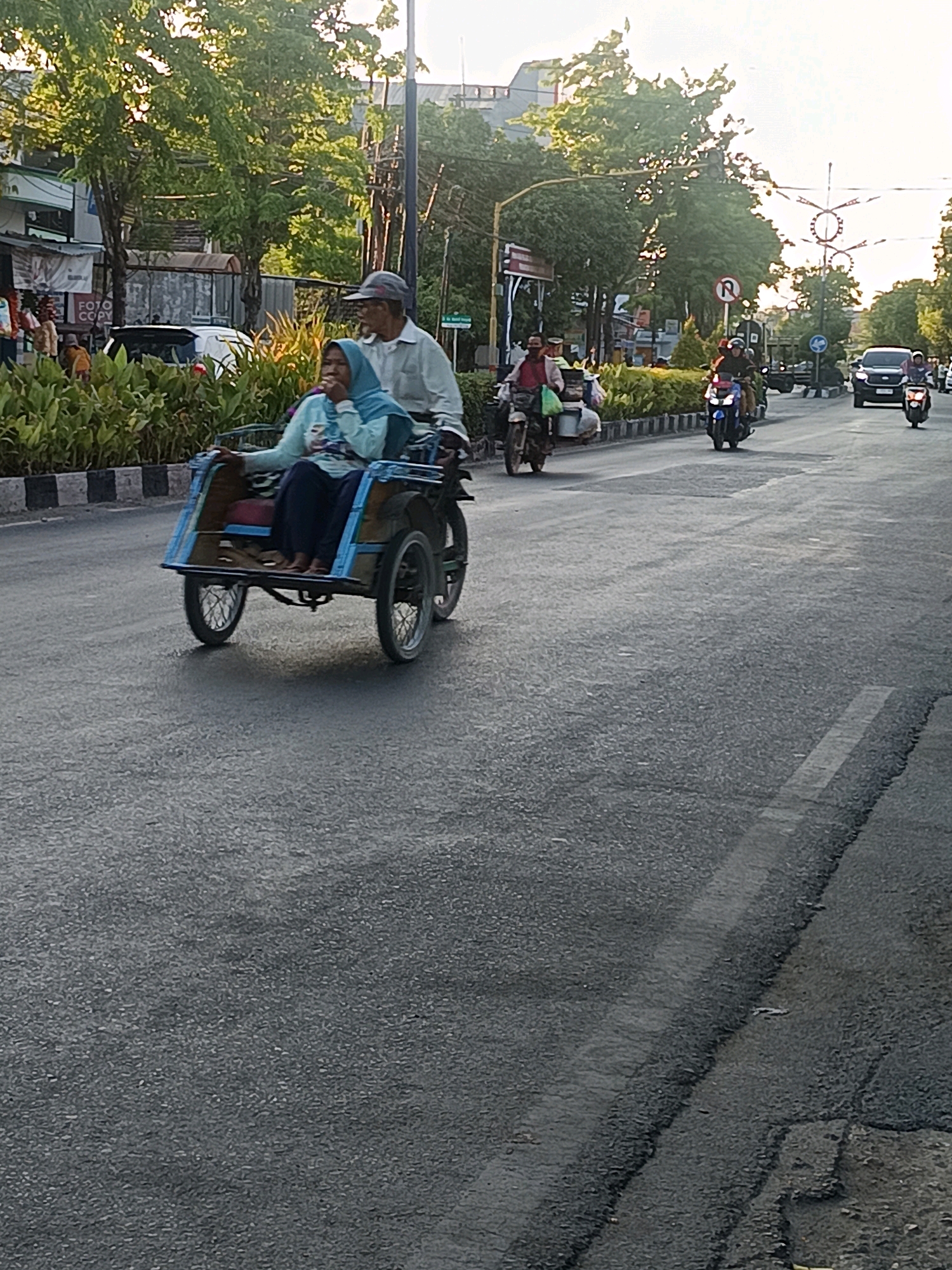A rickshaw, driven by a man wearing a cap and a light-colored shirt, is traveling down a street in the city of Gunung Sekar, Indonesia. A woman in a light blue hijab and matching top is seated in the passenger section of the rickshaw. The street is paved and appears to be wet, with some patched areas. Several other vehicles, including motorcycles and a car, are also visible on the road. The background features trees, buildings, and streetlights. The sky is bright, suggesting it is daytime.