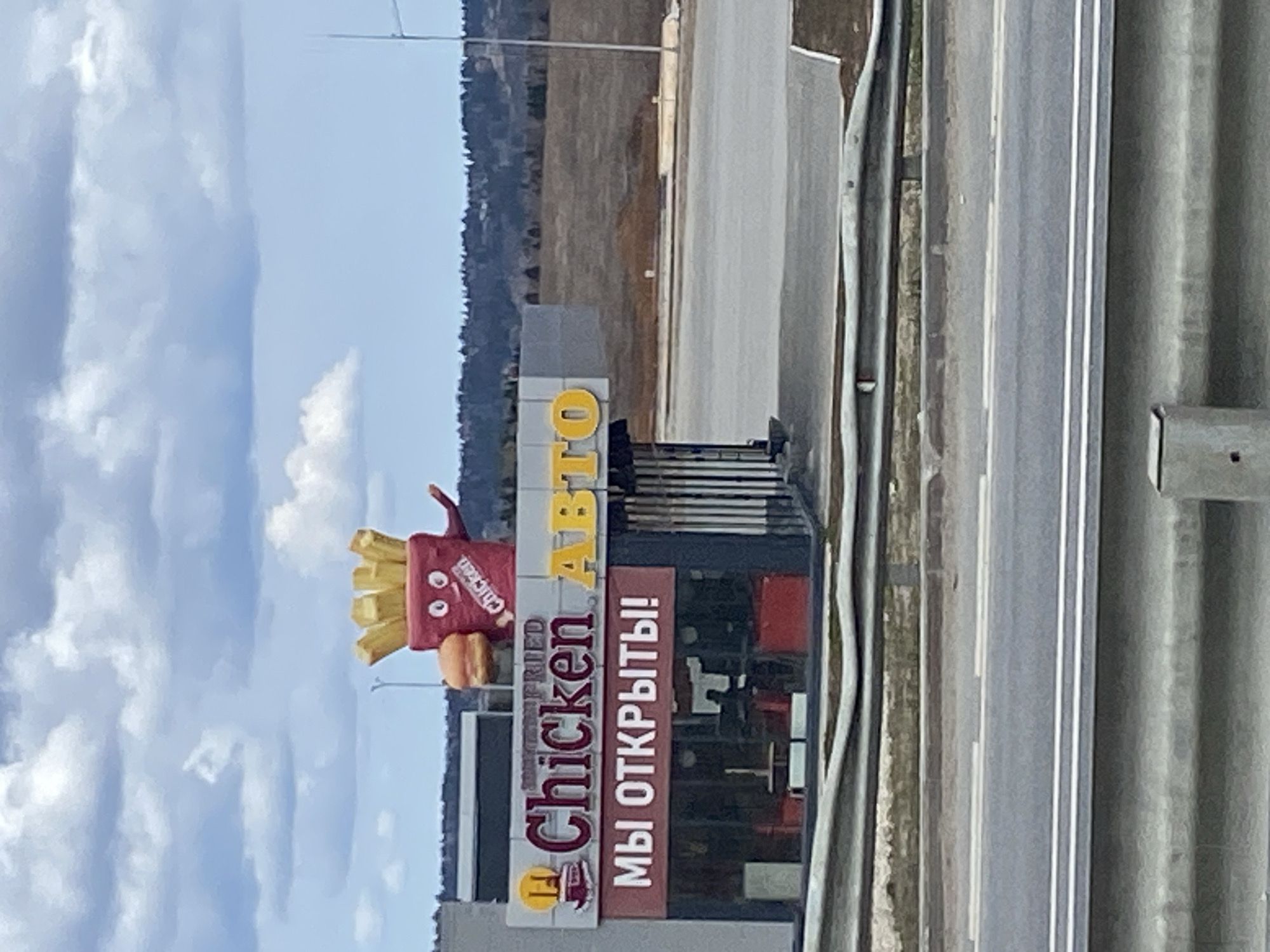 Here is a description of the image:
The image shows a restaurant sign. The sign features the words "Chicken Fried" stacked vertically, with an image of a chicken bucket above the word "Chicken" and a fry container with a face above the bucket. To the right of the "Chicken Fried" sign is a sign with the word "АВТО" above the phrase "МЫ ОТКРЫТЫ!". The sign is located next to a road and appears to be near a highway. The sky is visible in the background with some cloud cover. It is difficult to identify a specific location based solely on the image content.