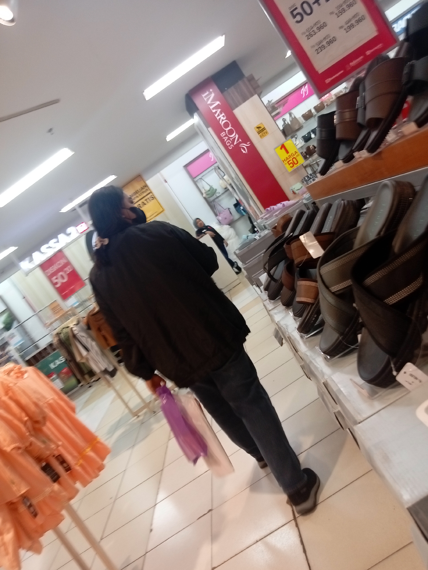 The image appears to be taken inside a retail store, likely a department store or mall, in Batam, Indonesia, given the prevalence of Indonesian signage and the typical store layout. The perspective is from a low angle, looking up and to the right.
In the foreground, a person is walking away from the camera, wearing a dark jacket and dark pants. They are carrying a purple shopping bag in their right hand and a white bag in their left. The flooring is tiled in a light beige color.
To the right of the person, there is a display of footwear, primarily sandals and flip-flops, arranged on shelves. These items are mostly dark in color, with some brown and black options visible. Price tags and branding are present on the footwear and shelves, though illegible due to the image quality and distance.
In the background, further displays of clothing and accessories are visible. To the left, there are racks of clothing, including some orange garments prominently displayed. Signs with Indonesian text are visible throughout the store. One sign on the left reads "KASSA2" and another red sign reads "DISKON 50%". Further in the background, a sign with "MAROON BAGS" is visible. Another sign with "HARGA 50%" indicates a sale.
The lighting appears to be bright, typical of indoor retail environments, with fluorescent lights visible on the ceiling. The overall impression is a busy commercial space with shoppers browsing merchandise.